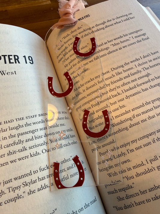 Detail shot of an aesthetic pink ribbon bookmark with horseshoes and stars held over an open Elsie Silver book. The rustic wooden background highlights the vibrant pink and clear acrylic design. This bookmark is designed for those who love "he falls first" tropes and southern belle aesthetics. A premium bookish accessory that is scratch-resistant. Perfect for organizing a home library or as a treat for yourself to celebrate a new romance book haul or Kindle accessory