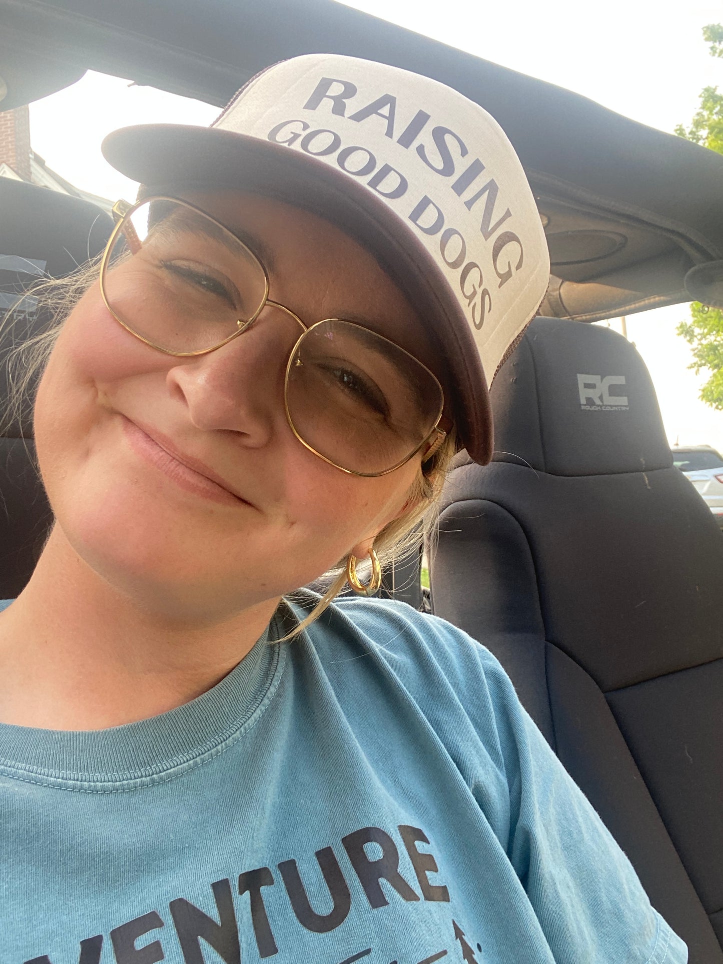 Person wearing a Raising Good Dogs brown and tan trucker hat inside a vehicle