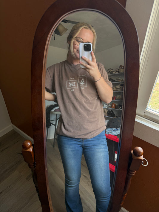 Person taking a mirror selfie wearing a brown t-shirt with tan Blessed with the Best text and graphic of two stamps with dogs on them and blue jeans in a room.
