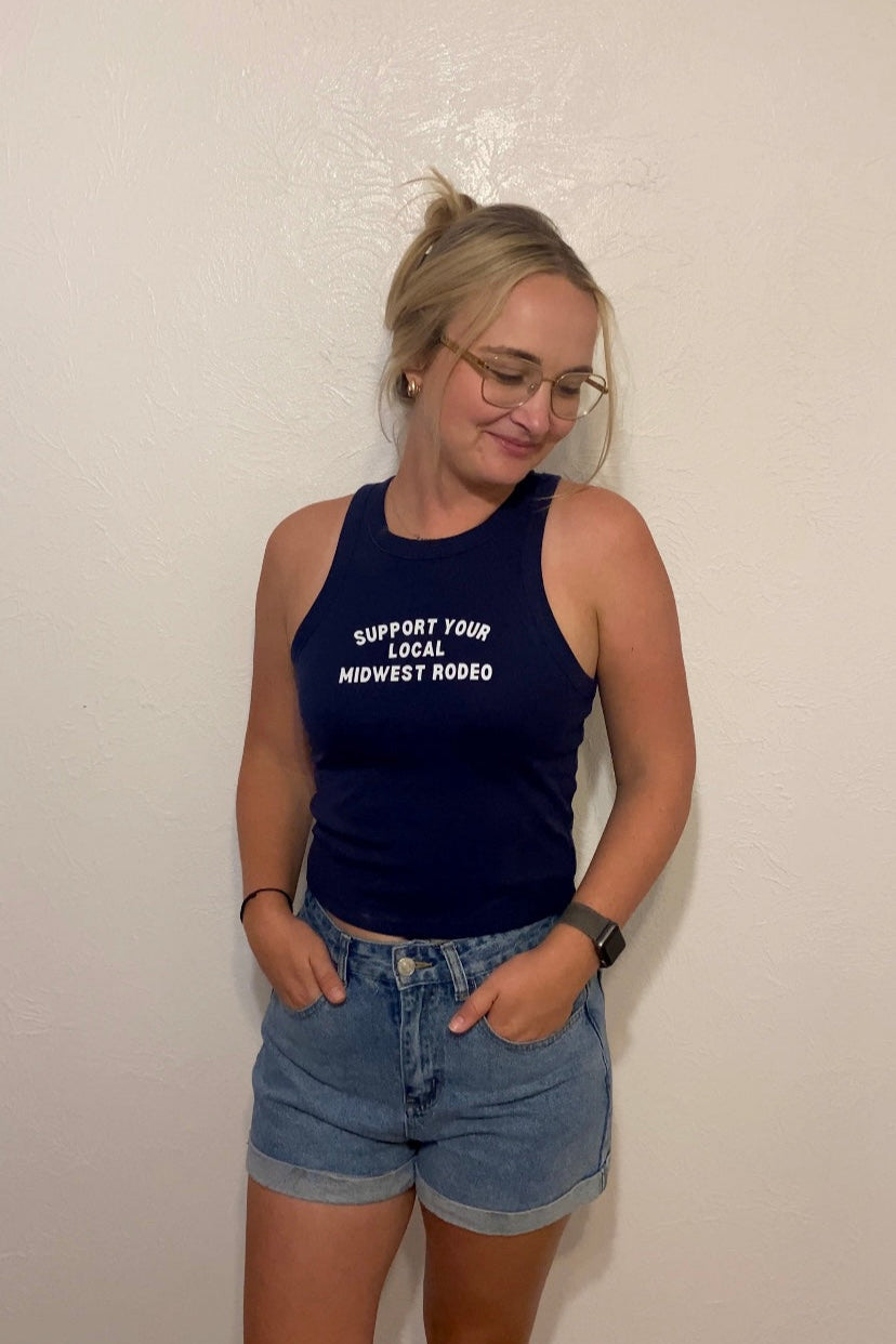 Woman wearing a navy tank top with white text that says "Support Your Local Midwest Rodeo" and denim shorts against a plain background