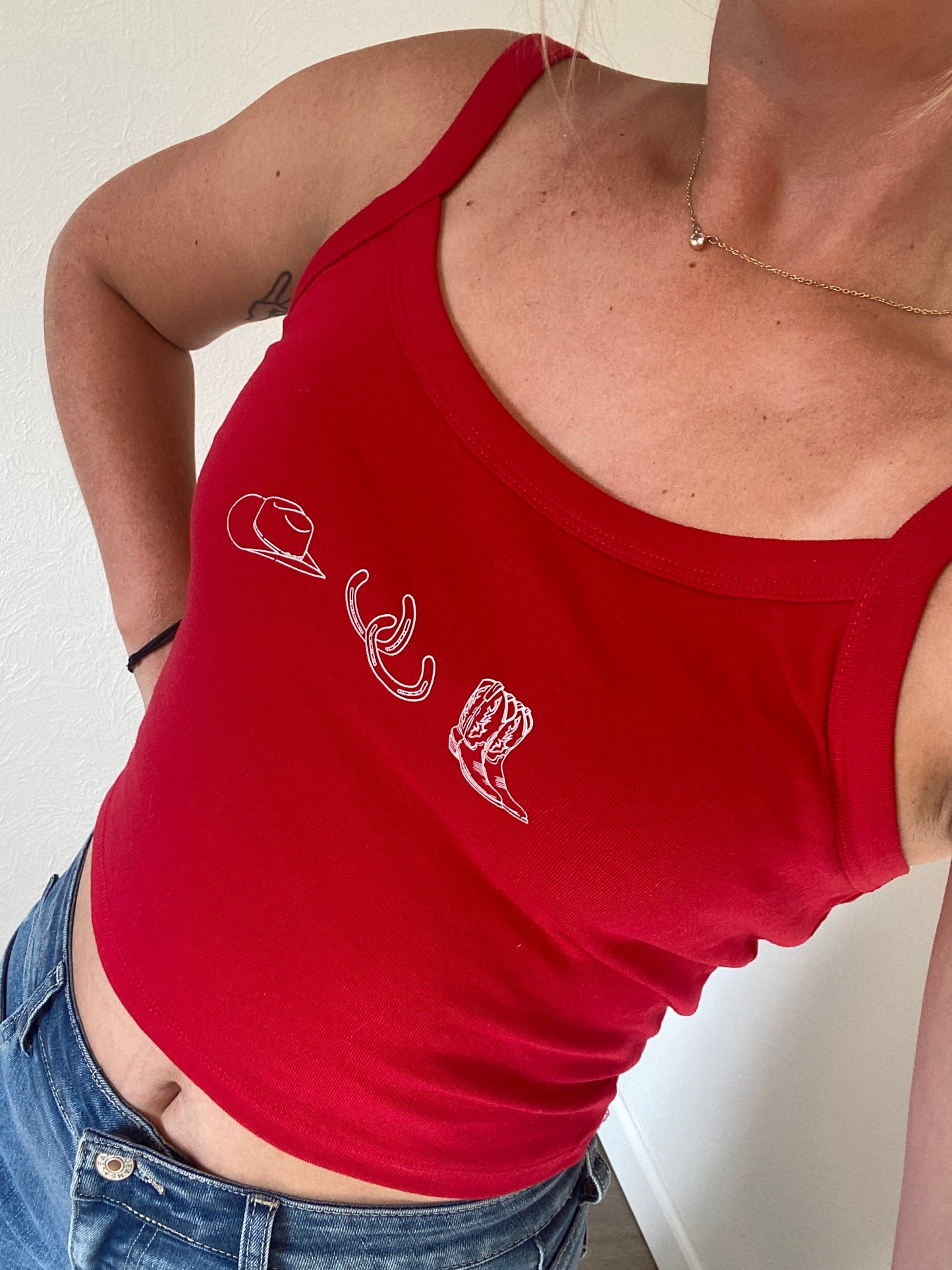 Person wearing a red cowgirl collage tank top with white graphics of a cowboy hat, horseshoes, and cowgirl boots on a neutral background with jean shorts