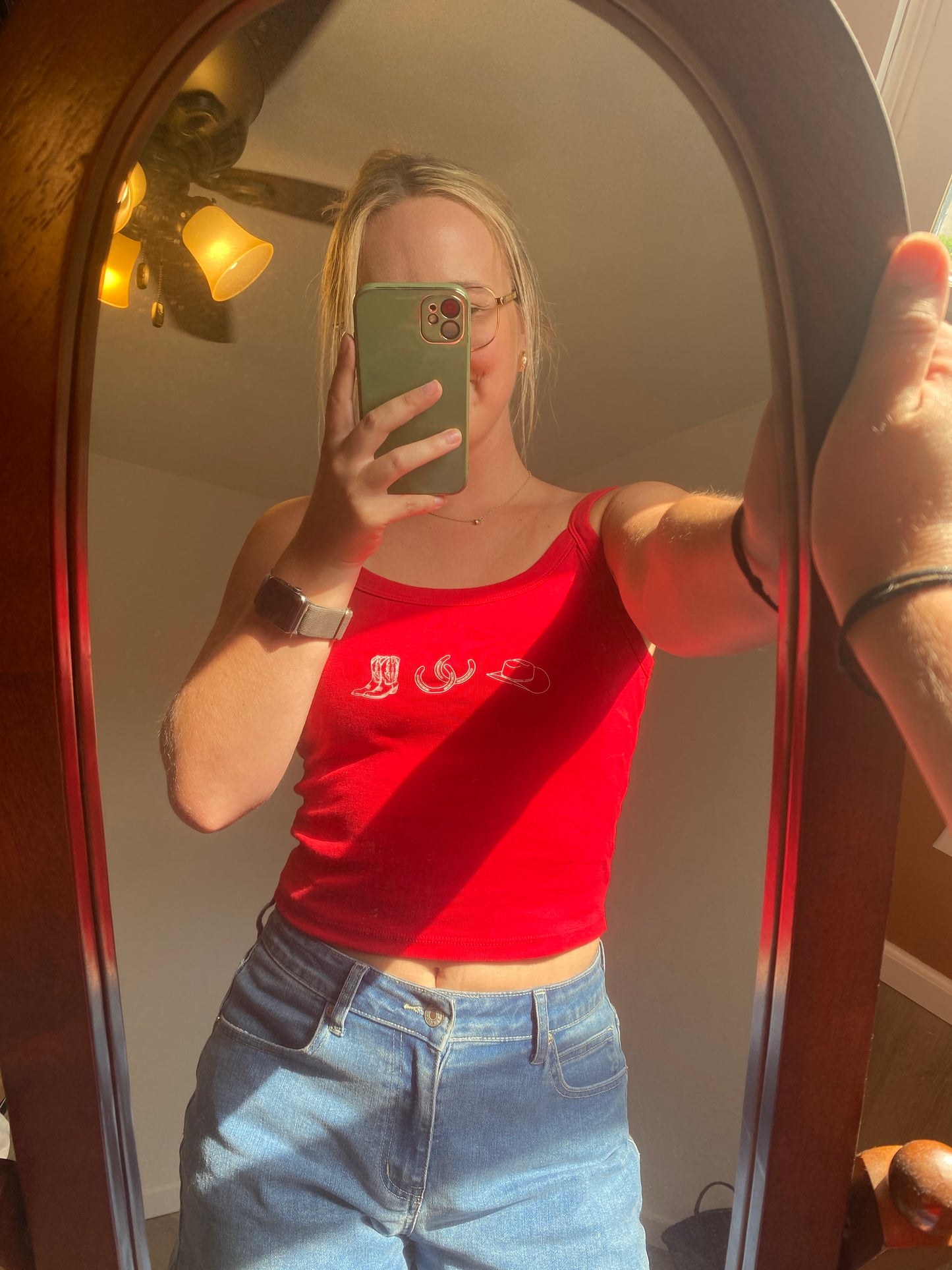 Person taking a mirror selfie wearing a red cropped tank shirt with cowboy boot, horseshoe, and cowgirl hat graphics and blue jeans.