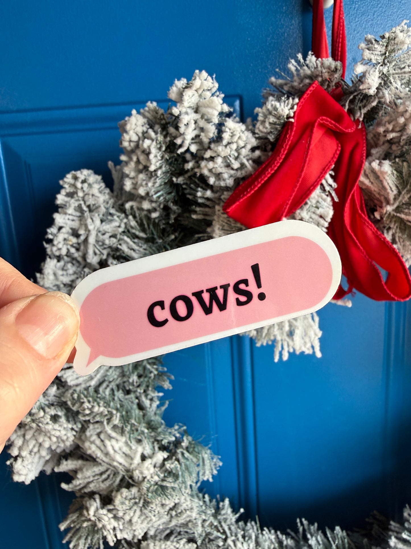 A "Cows!" pink text bubble sticker held up outdoors in front of a blurred blue front door with a decorative wreath, showcasing the sticker's colors in natural bright daylight.
