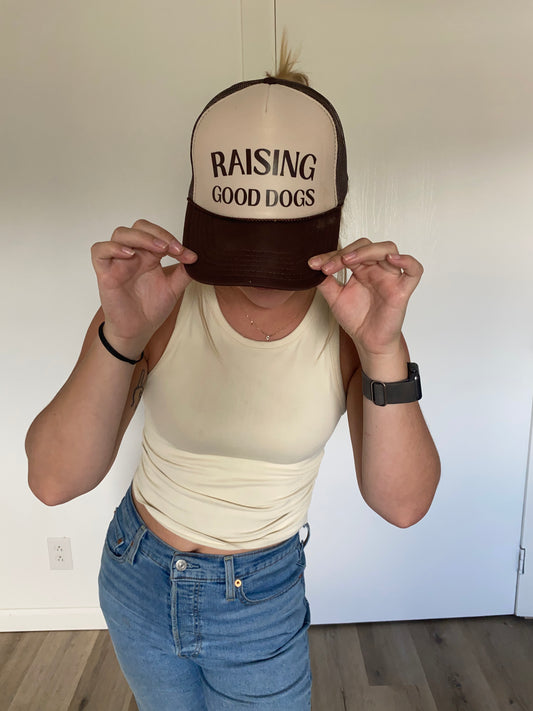 Person wearing a two tone, brown and tan cap with 'Raising Good Dogs' text on a white background