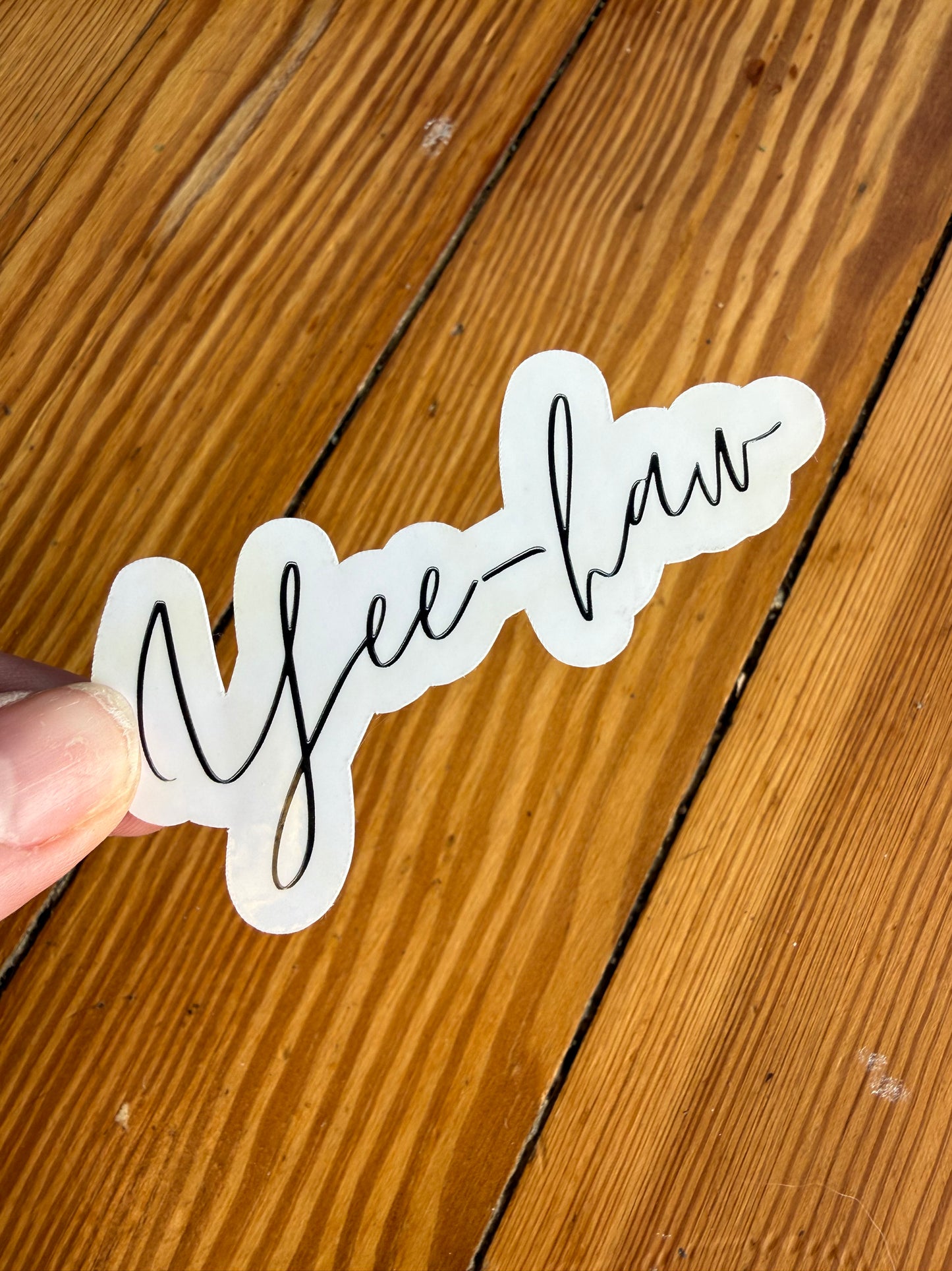 A hand holding a 3-inch "Yee-haw" cursive vinyl sticker against a light wood background, showing the light reflecting off the high-shine glossy finish.