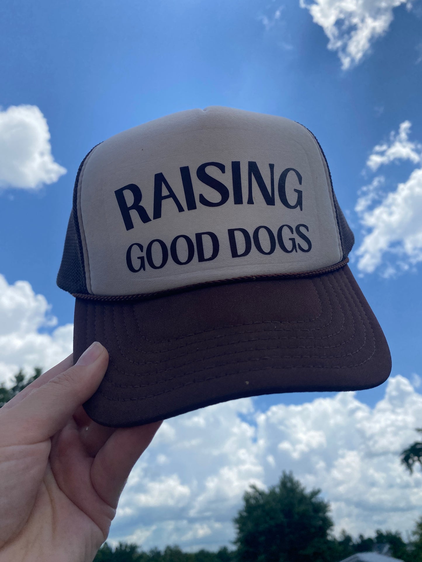 Brown and Tan Foam Trucker Hat with 'Raising Good Dogs' in brown text held against a blue sky with clouds