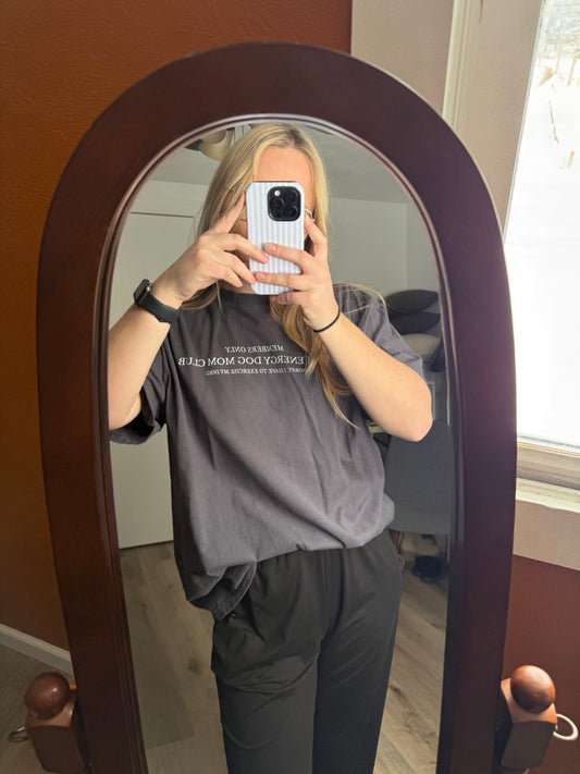 A lifestyle mirror selfie of a woman wearing an oversized dark gray High Energy Dog Mom Club t-shirt paired with casual black sweatpants. She is holding her phone with both hands, showing off the "Members Only" and "Sorry I Have to Exercise My Dog" text. This trendy dog mom outfit is ideal for walking the dog, going to the park, or running errands. The oversized fit provides a cozy, athletic aesthetic for women who prioritize their pet's active lifestyle and daily exercise.