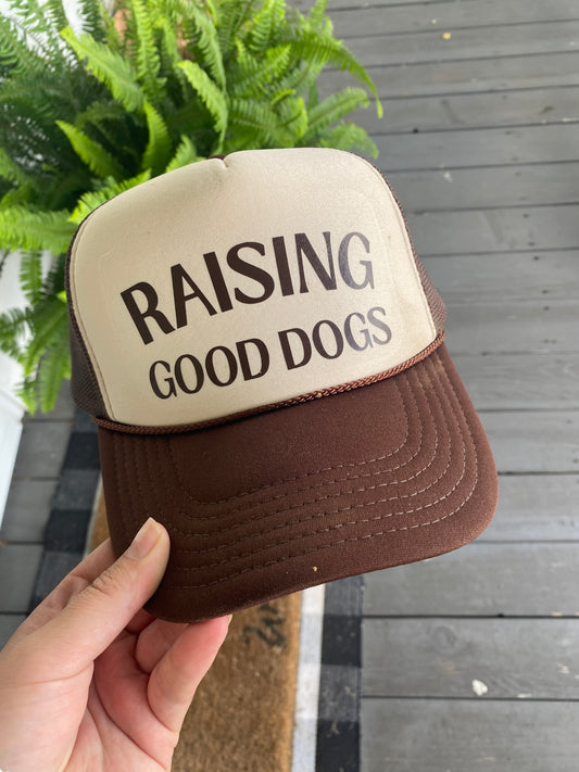Brown and beige trucker hat with 'Raising Good Dogs' text held by a hand against a wooden door background.