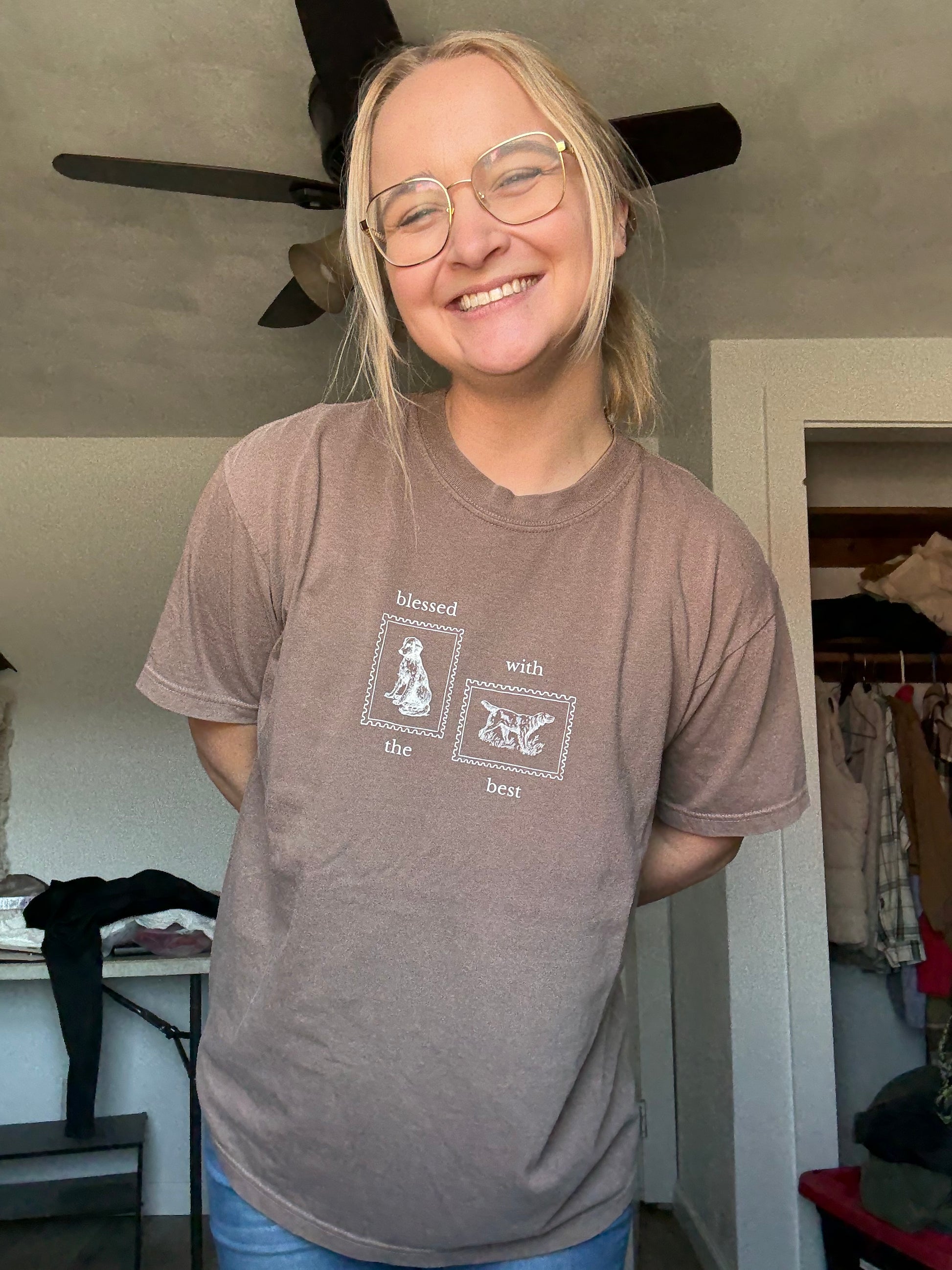 Person wearing a brown t-shirt with tan Blessed with the Best text and graphic of two stamps with dogs on them, standing indoors.