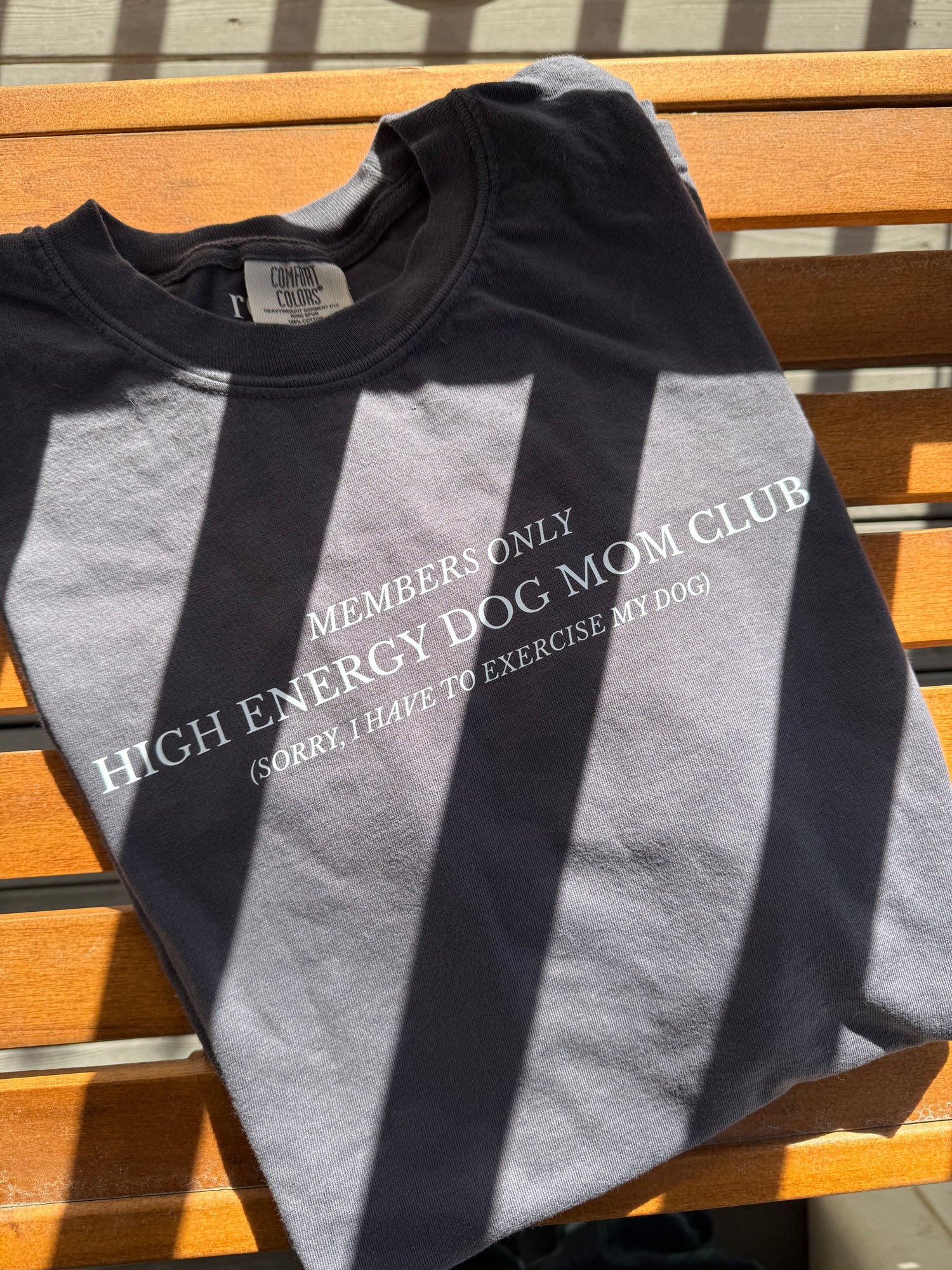 A flat lay photograph of a dark gray Comfort Colors 1717 t-shirt folded neatly on a rustic wooden bench in bright, natural morning sunlight. The shirt features a white graphic design that reads "High Energy Dog Mom Club, Members Only, Sorry I Have to Exercise My Dog." This high-quality cotton garment is perfect for active pet owners, aussie moms, border collie owners, and fitness enthusiasts. The aesthetic highlights the soft, vintage-wash fabric texture and durable screen print.
