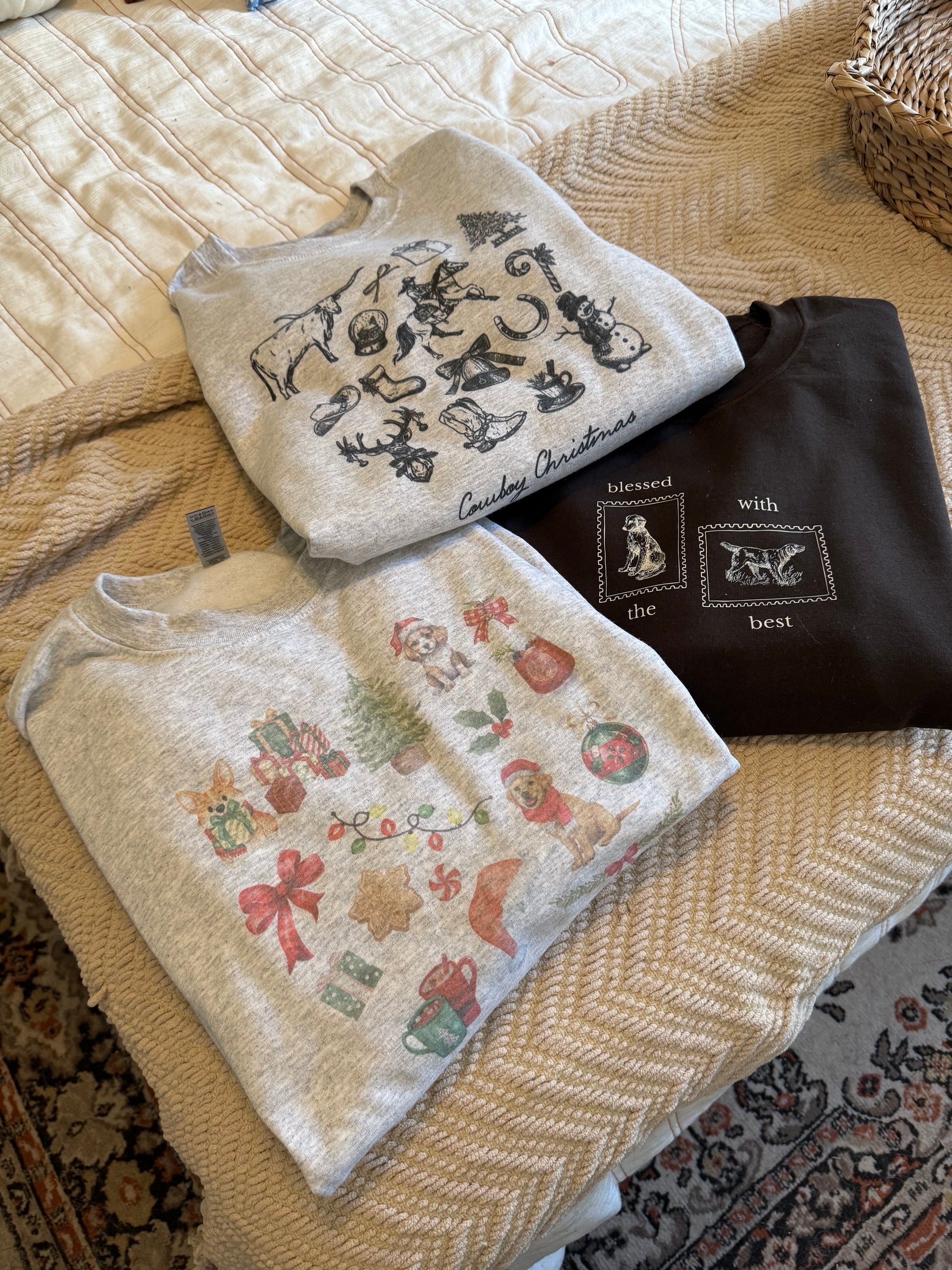 Ash gray Gildan 18000 watercolor dog Christmas sweatshirt folded on neutral bedding alongside cowboy Christmas sweatshirt and Thanksgiving crewneck, holiday gift ideas, Western Christmas style, cozy seasonal unisex sweatshirts for pet lovers and festive family outfits.