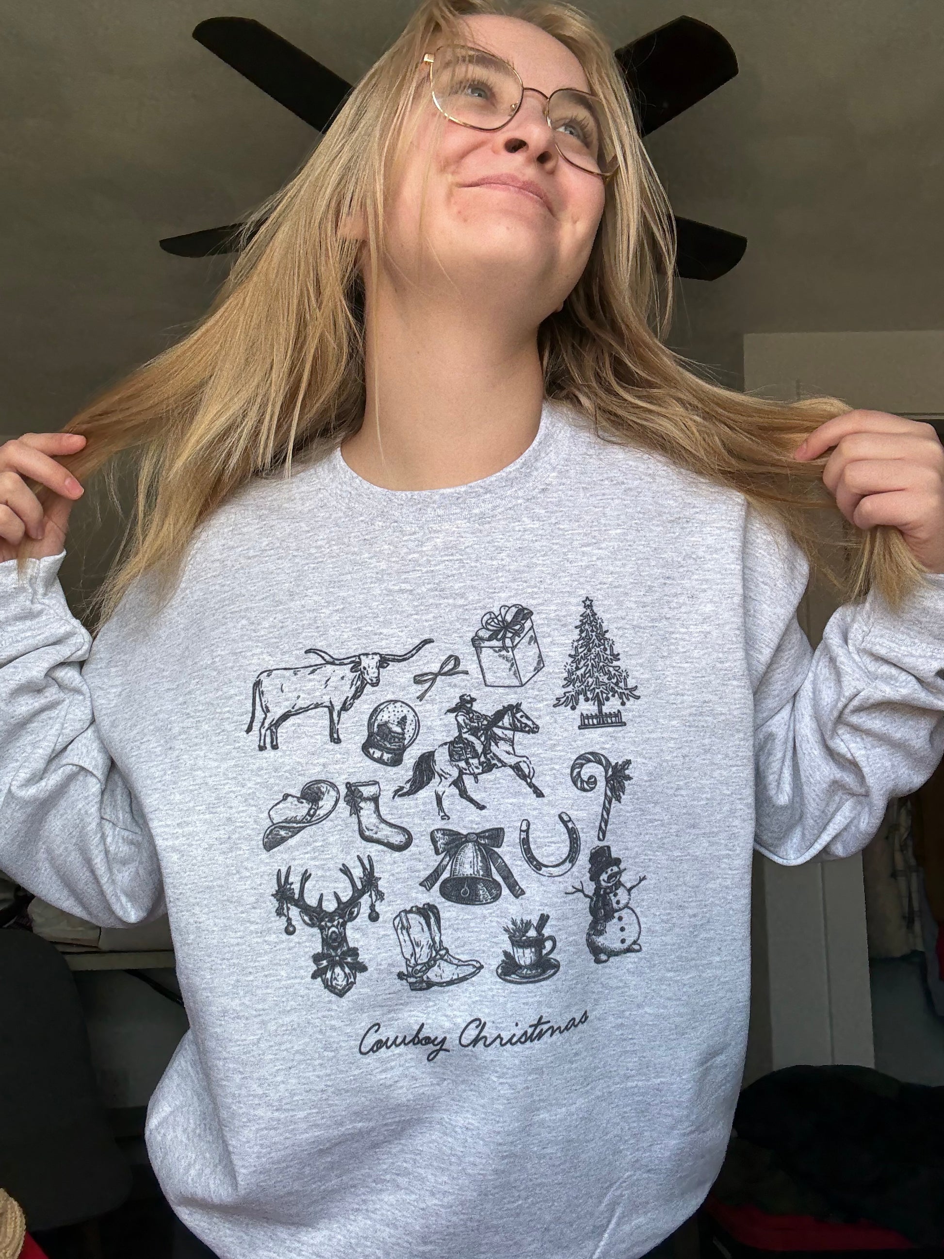 A woman lifting her hair to show the ash-colored cowboy Christmas crewneck featuring a detailed vintage Western holiday collage of cowboy illustrations, Christmas sketches, boots, hats, and festive retro accents. Long-tail keywords for rustic cowgirl Christmas gifts, Western holiday outfit ideas, cozy winter cowgirl crewnecks, and aesthetic retro Christmas cowboy apparel.