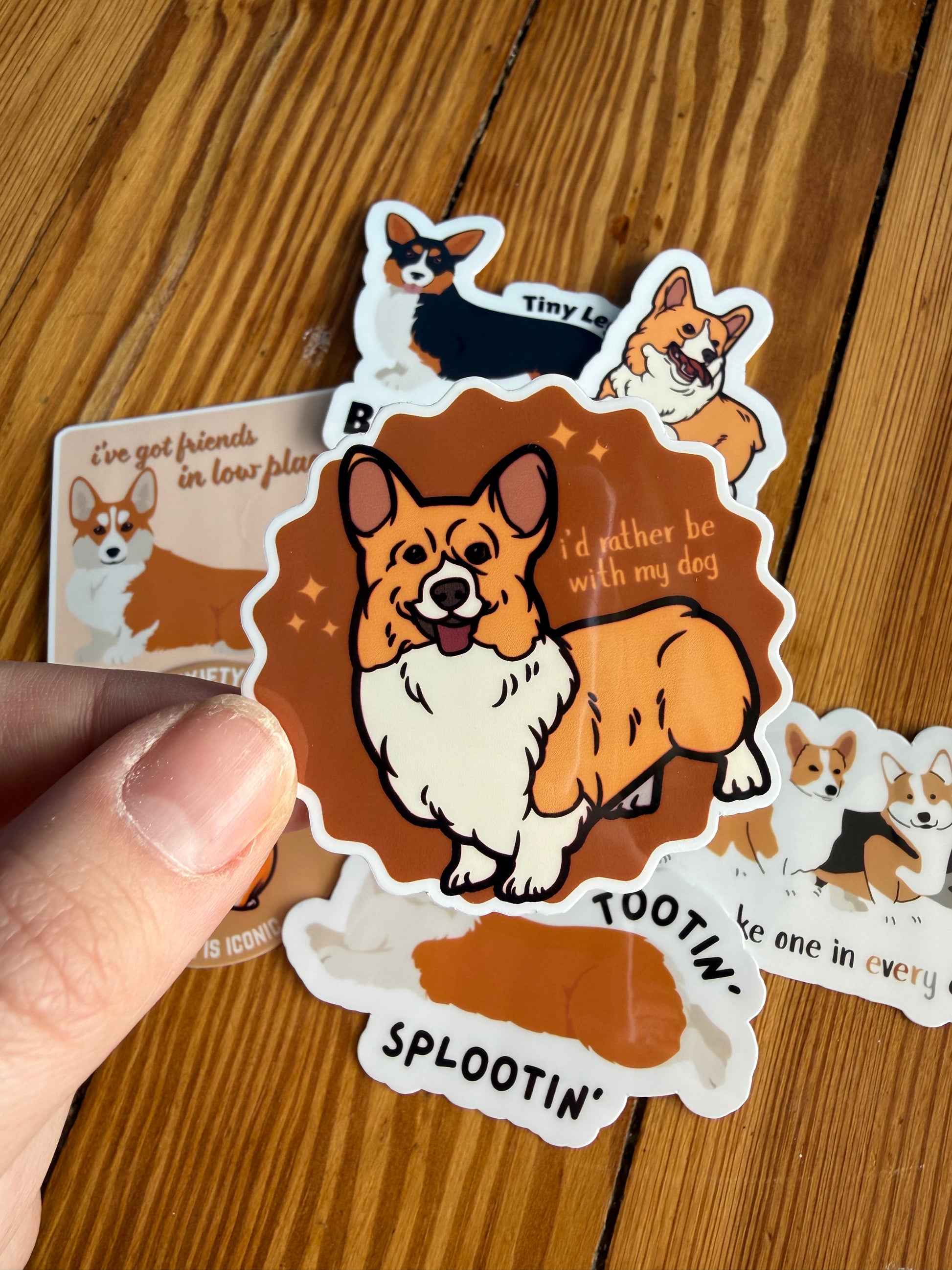 "I'd rather be with my dog": Close-up of a sticker held over a wooden floor featuring a cute corgi illustration and the text "I’d rather be with my dog."