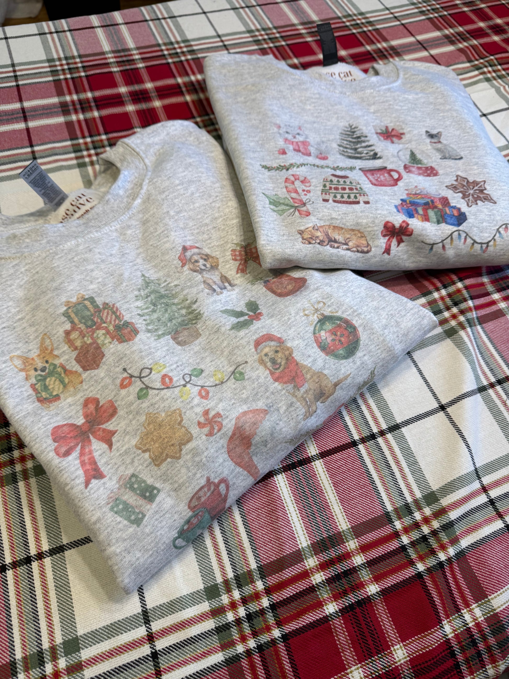 Folded Gildan 18000 ash Christmas dog sweatshirt next to matching cat Christmas design on red plaid background, cozy pet lover holiday apparel, watercolor dog and cat illustrations, aesthetic gift ideas for dog moms and cat moms during Christmas season.