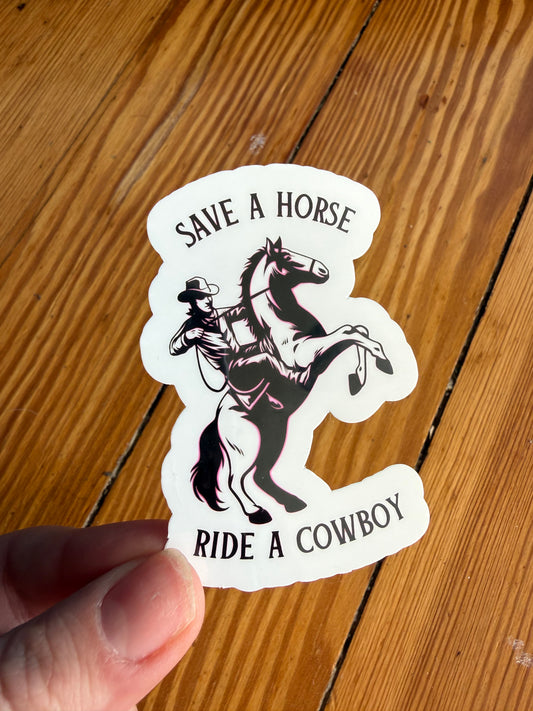 Hand holding a western cowboy sticker featuring a cowboy on a rearing horse with the phrase “Save a Horse, Ride a Cowboy.” This country western vinyl sticker is shown against a rustic wooden background, highlighting its size and detail. Perfect for cowboy lovers, western humor fans, country girls, rodeo enthusiasts, and anyone who loves funny western decals for laptops, tumblers, journals, and phone cases.
