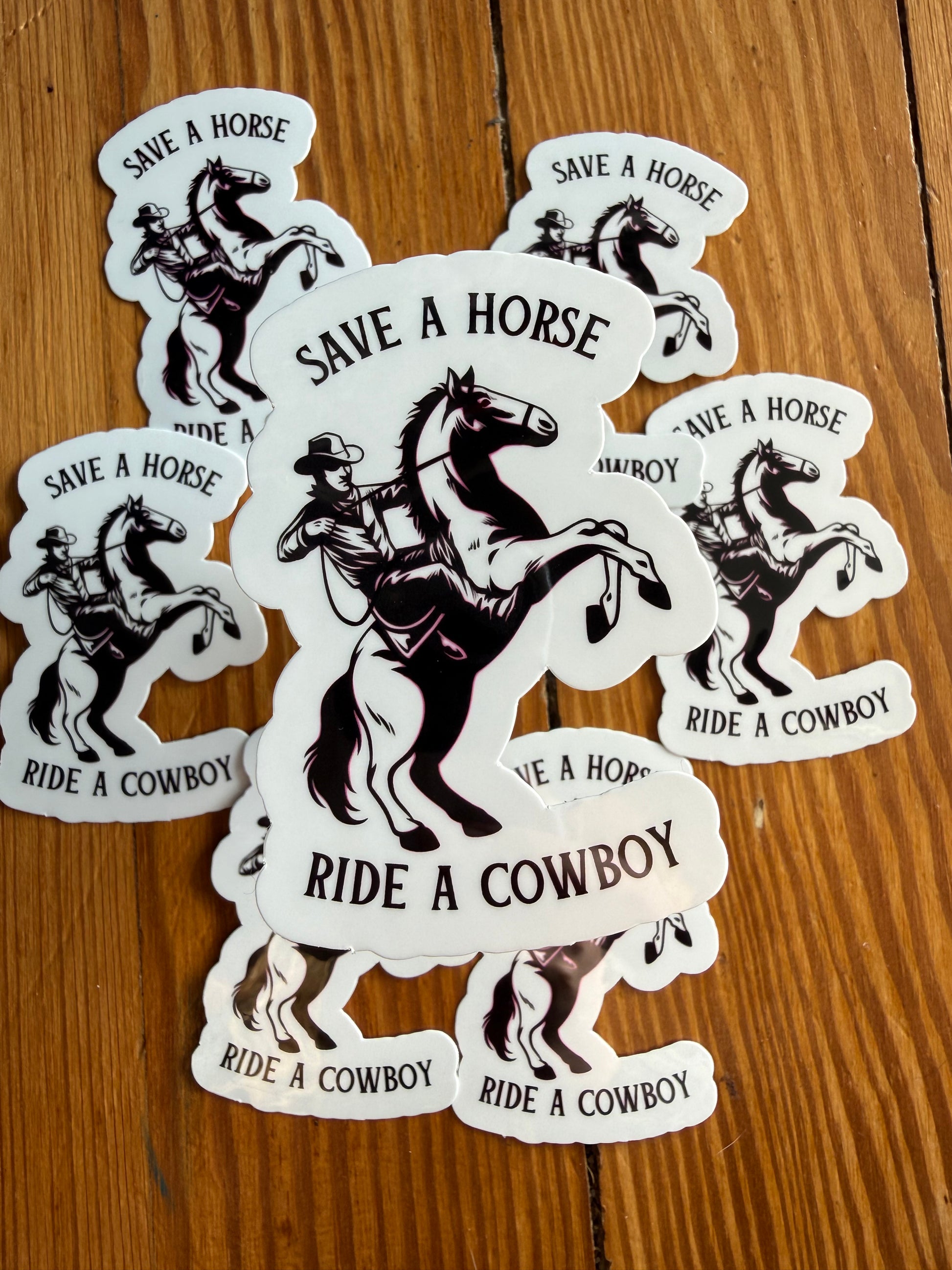 Multiple “Save a Horse, Ride a Cowboy” stickers displayed on a rustic wooden background, with one sticker hovering above the others to show thickness and quality. This western cowboy sticker features a bold cowboy on a rearing horse and a fun country saying. Ideal as a funny western sticker, cowboy aesthetic decal, rodeo sticker, country music gift, or vinyl sticker for laptops, notebooks, guitars, and gifts.