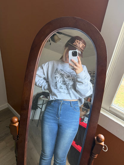 A woman taking a mirror selfie while wearing an ash cowboy Christmas sweatshirt with a vintage Western holiday collage, paired with jeans and a brown COWGRL trucker hat. Retro cowboy sketches and festive Christmas art create a rustic cowgirl winter look. Long-tail keywords include Western Christmas crewneck, cowgirl aesthetic holiday outfit, rodeo Christmas gifts, and vintage cowboy Christmas apparel.