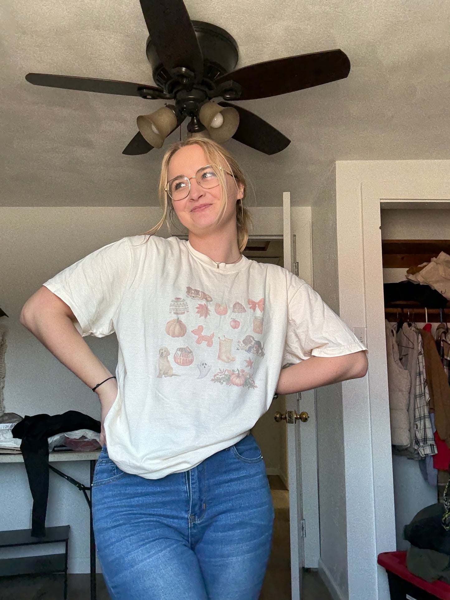 Lifestyle photo of woman wearing sublimated fall dog collage Comfort Colors ivory tee paired with jeans. Autumn dog mom graphic shirt with watercolor pumpkins, dogs, leaves, and cozy fall accents. Trendy fall outfit idea for pet owners, dog lovers, and fans of vintage aesthetic seasonal shirts.