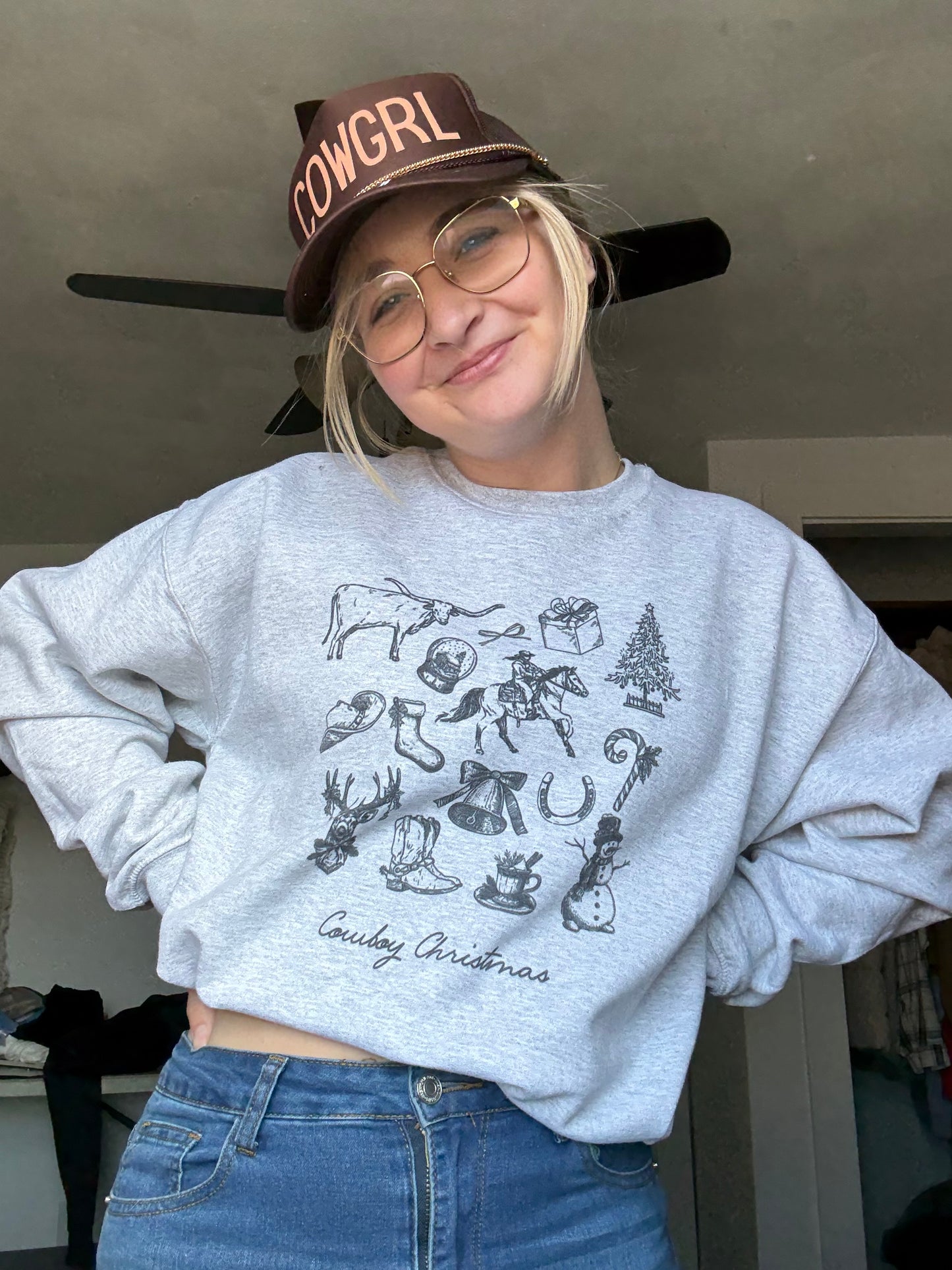 A woman wearing an ash cowboy Christmas crewneck with a vintage Western holiday collage, styled with jeans and a brown COWGRL trucker hat. Retro cowboy sketches and Christmas illustrations create a rustic cowgirl holiday aesthetic. Perfect long-tail keywords for Western Christmas outfits, cowgirl winter apparel, rodeo Christmas gift ideas, and rustic farmhouse holiday clothing inspiration.