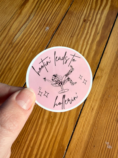 Hand holding a 2-inch round pink vinyl sticker that says “Hootin’ Leads to Hollerin’,” displayed against a rustic wooden background. This funny southern saying sticker features playful country humor and a bold pink design. Perfect for cowgirls, country music lovers, southern humor fans, and western aesthetic lovers looking for a cute vinyl sticker for laptops, phone cases, planners, and journals.
