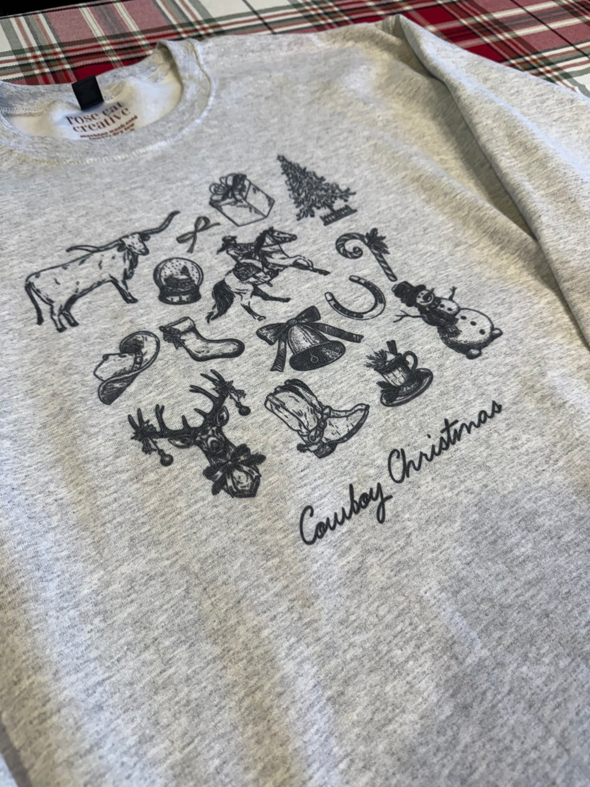 Ash gray cowboy Christmas crewneck laid flat on a red plaid background, showing a collage of vintage cowboy sketches mixed with classic Christmas drawings, Western boots, rope, stars, and retro festive art. Perfect long-tail keywords for rustic Western Christmas home decor photos, vintage cowgirl holiday clothing, and farmhouse Christmas outfit inspiration.