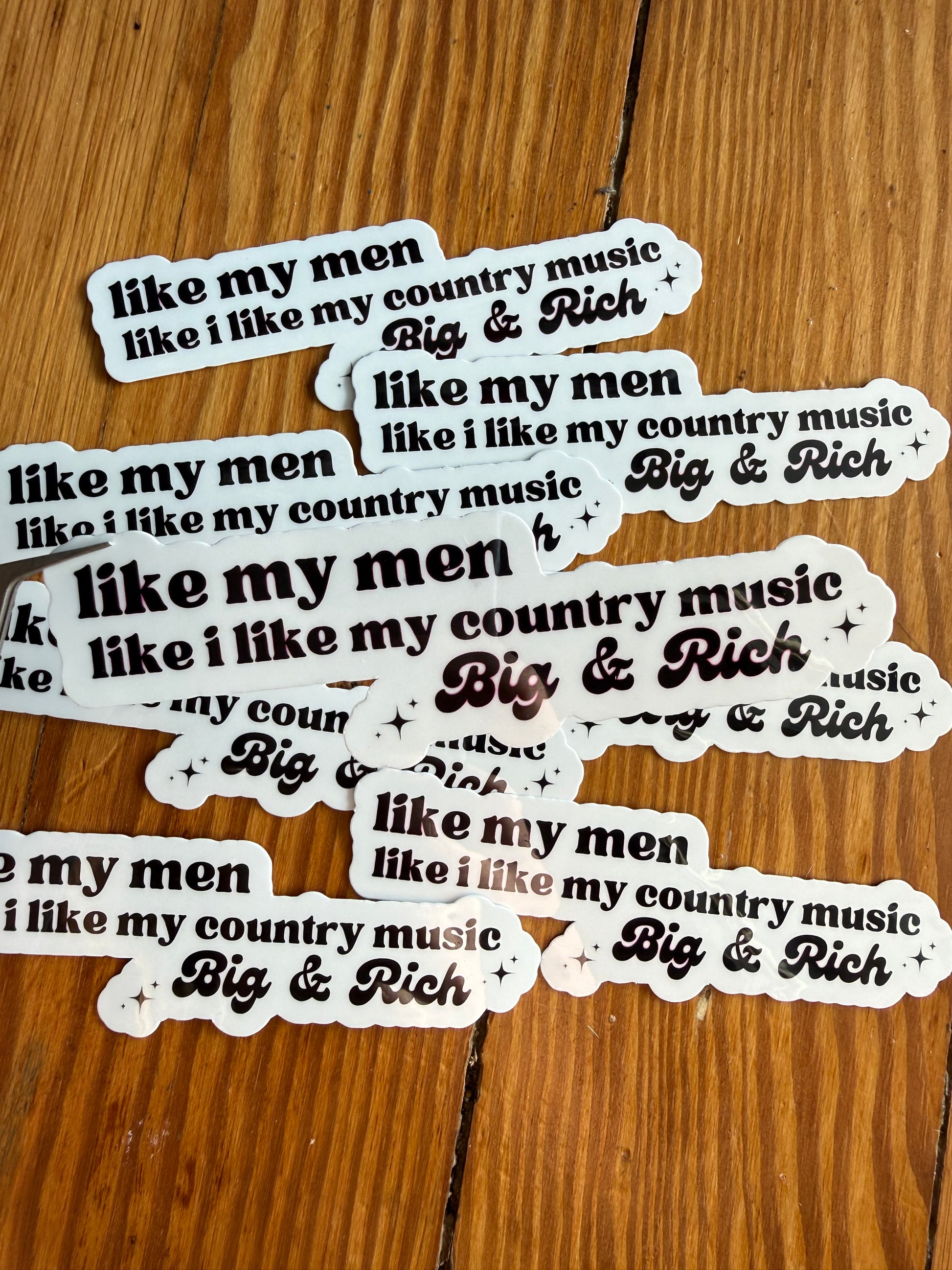 Single vinyl country music sticker featuring the quote “I like my men like I like my country music, Big & Rich,” shown hovering over a background of printed photos. This humorous country lyric inspired sticker is ideal for fans of Big & Rich, southern sayings, cowgirl humor, country concert merch, car decals, planner stickers, and unique country themed gifts for women and men.