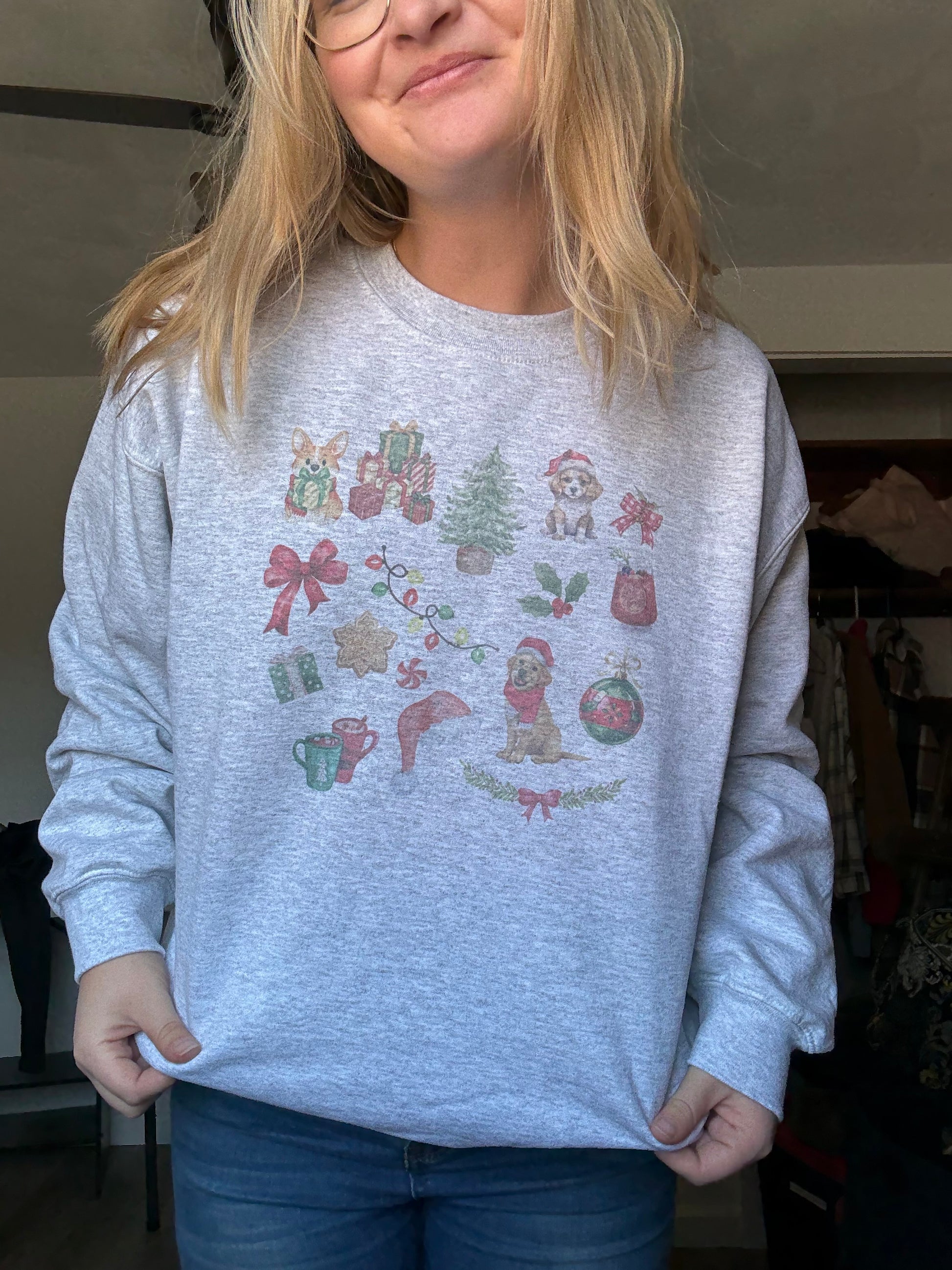 Person wearing and holding up Gildan 18000 ash gray Christmas dog sweatshirt with watercolor collage of holiday dog illustrations, cozy unisex crewneck winter outfit, trendy dog lover Christmas apparel, soft fleece gift for pet owners, aesthetic holiday sweatshirt worn with casual jeans.