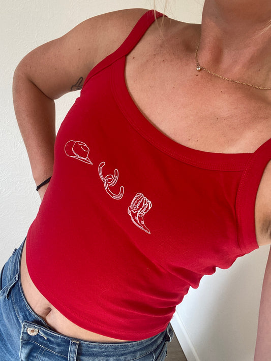 Person wearing a red cowgirl collage tank top with white graphics of a cowboy hat, horseshoes, and cowgirl boots on a neutral background with jean shorts