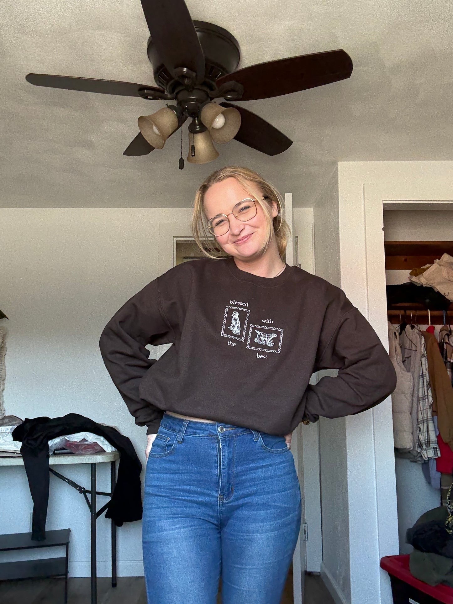 Person wearing a dark brown dog lover sweatshirt with Blessed with the Best text and dog stamp graphics and jeans in a room with a ceiling fan.