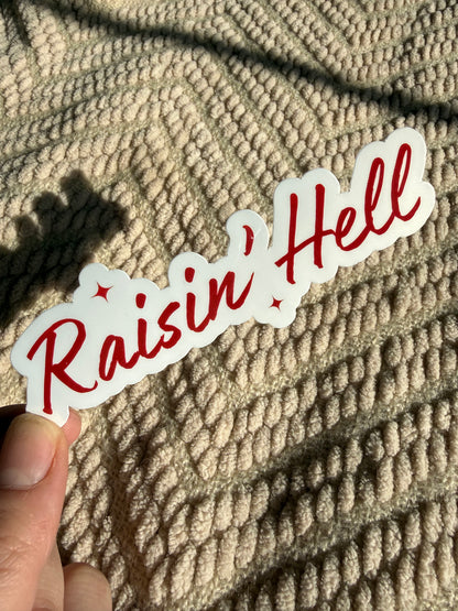 Close-up of "Raisin’ Hell" sticker in red cursive font; edgy southern style decal for car windows and water bottles, glossy finish with sunlit background.