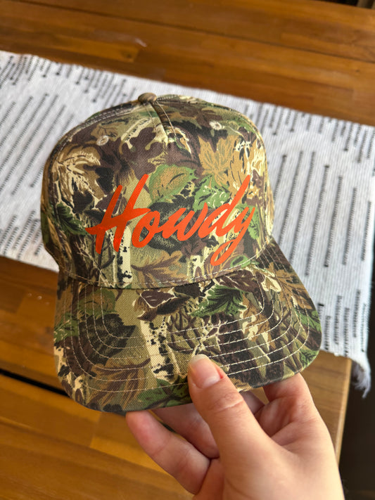A close-up view of a camo snapback hat featuring bright orange "Howdy" script lettering, held in front of a modern farmhouse table with a black and white patterned runner. The focus is on the high-quality embroidery and classic forest camouflage pattern.