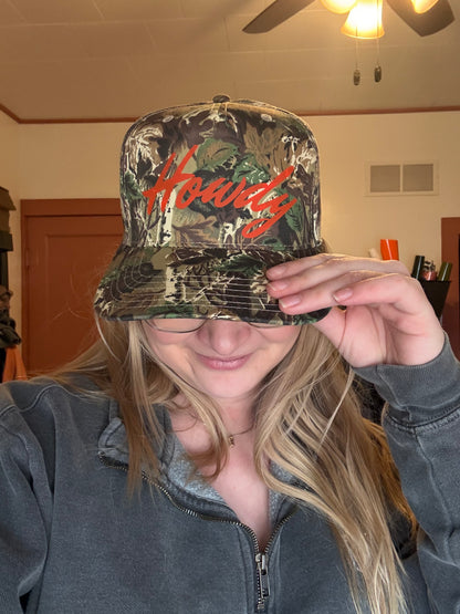 A young woman wearing a camouflage trucker hat with orange cursive "Howdy" embroidery, paired with a gray quarter-zip pullover. She is looking down with her hand resting on the hat bill, showing a trendy western streetwear aesthetic for fall or winter.