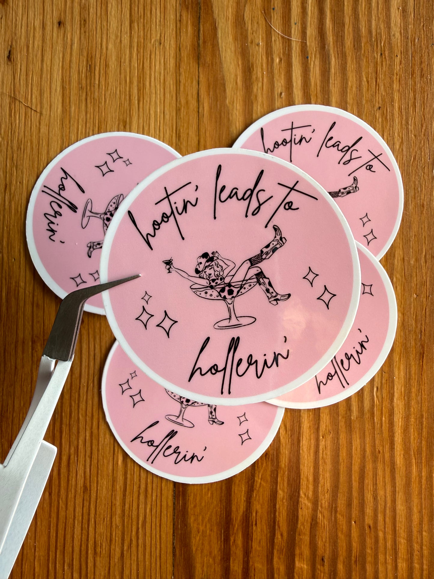A grouping of 2-inch round pink “Hootin’ Leads to Hollerin’” vinyl stickers displayed on a rustic wooden background, with one sticker held closer to the camera to show detail and finish. This funny southern saying sticker is ideal for cowgirl gifts, country humor lovers, western aesthetic collections, rodeo fans, bachelorette party favors, and vinyl decals for journals, laptops, and tumblers.