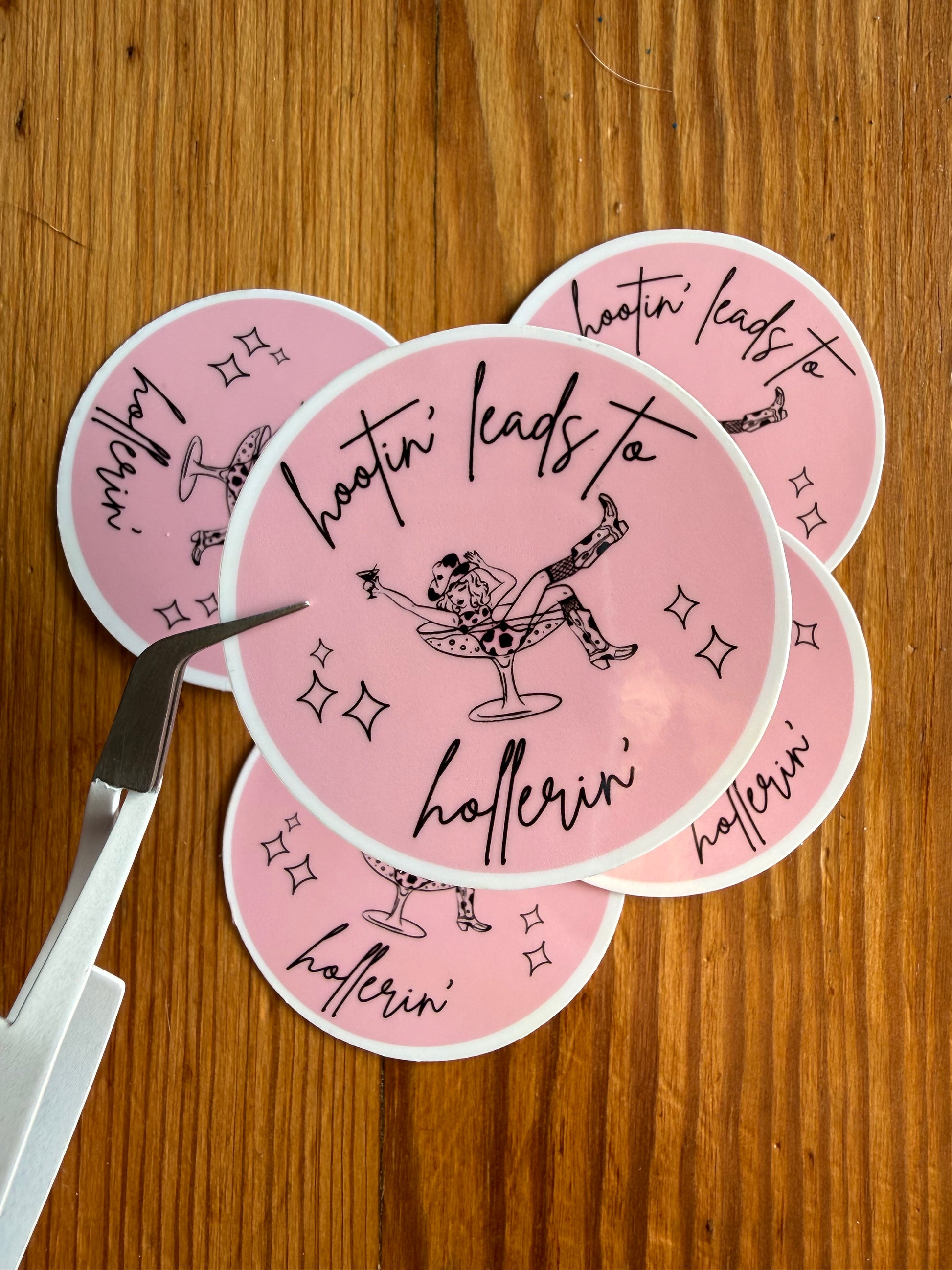 A grouping of 2-inch round pink “Hootin’ Leads to Hollerin’” vinyl stickers displayed on a rustic wooden background, with one sticker held closer to the camera to show detail and finish. This funny southern saying sticker is ideal for cowgirl gifts, country humor lovers, western aesthetic collections, rodeo fans, bachelorette party favors, and vinyl decals for journals, laptops, and tumblers.