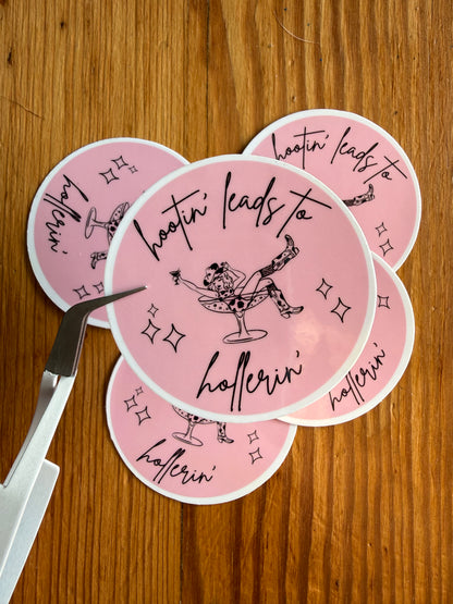 A grouping of 2-inch round pink “Hootin’ Leads to Hollerin’” vinyl stickers displayed on a rustic wooden background, with one sticker held closer to the camera to show detail and finish. This funny southern saying sticker is ideal for cowgirl gifts, country humor lovers, western aesthetic collections, rodeo fans, bachelorette party favors, and vinyl decals for journals, laptops, and tumblers.
