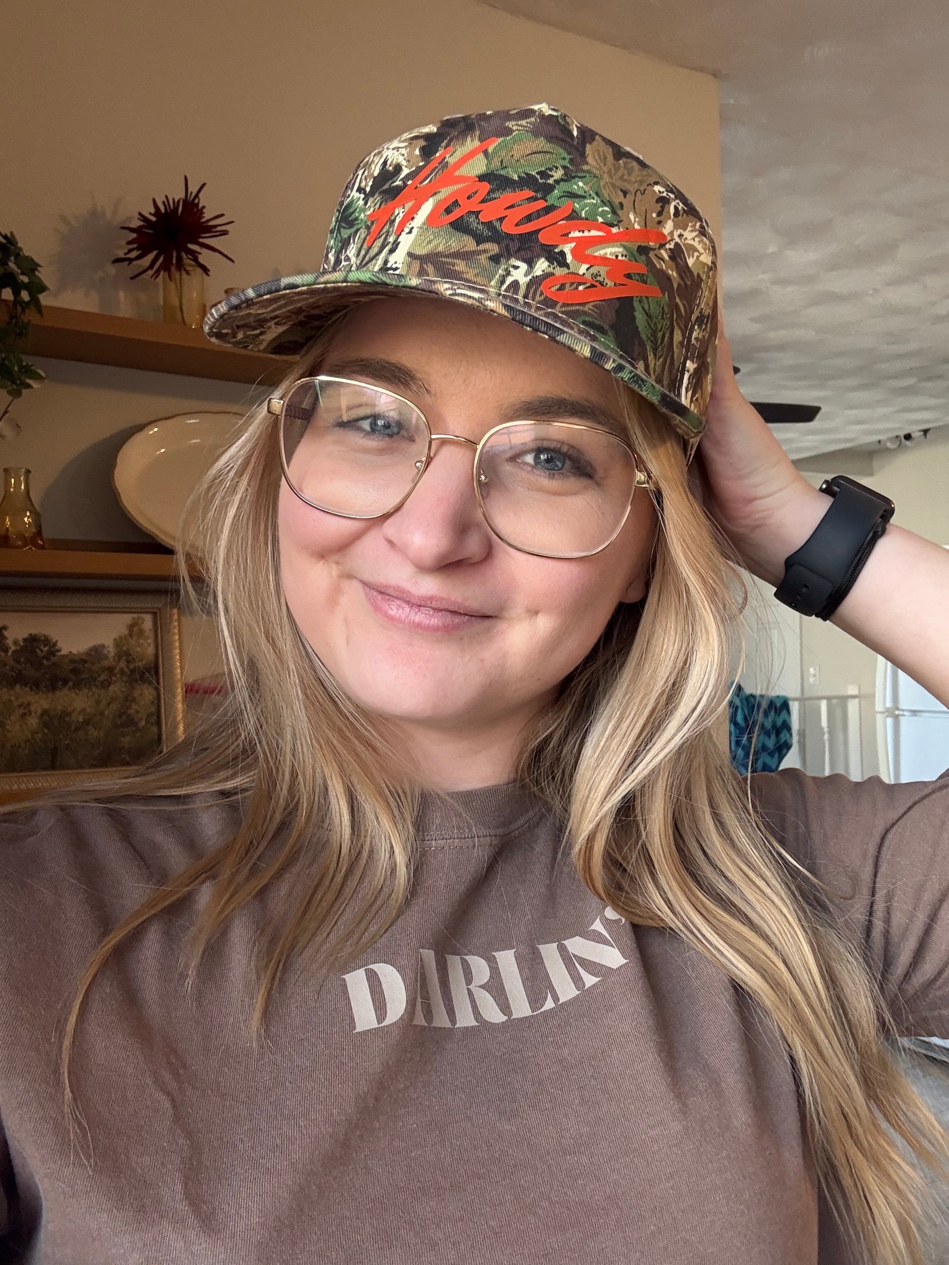 A smiling girl wearing a "Howdy" camo hat and a brown "Darlin'" graphic t-shirt. She is looking at the camera with her hand on the back of the hat, showcasing a cute coastal cowgirl outfit and southern style accessories for women.