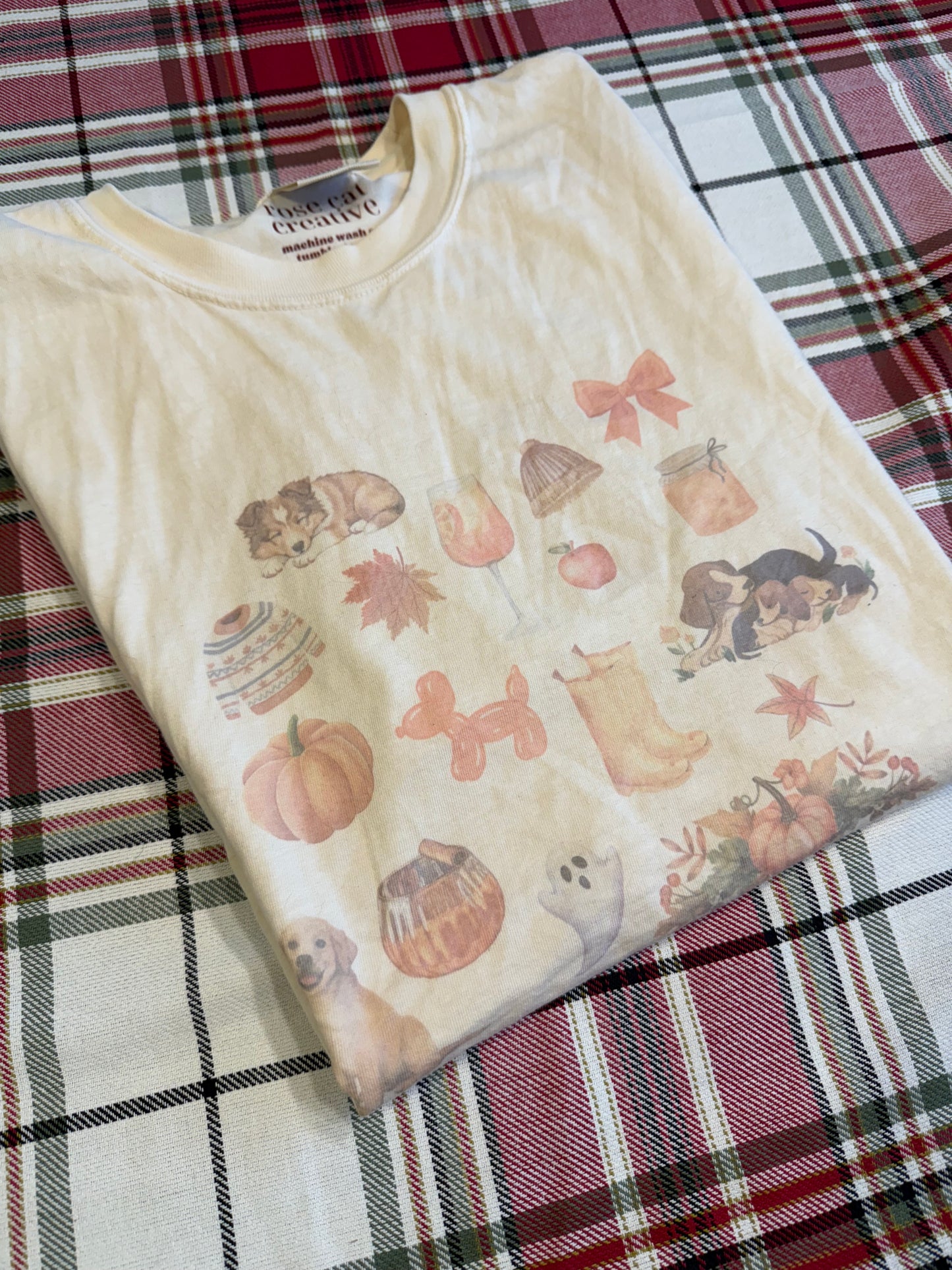 Flat lay of Comfort Colors ivory fall dog collage t-shirt on red plaid blanket. Sublimated watercolor fall and dog themed graphic tee for pet lovers. Cozy autumn apparel photography with rustic farmhouse fall accents. Perfect fall dog mom aesthetic shirt for gift giving, Thanksgiving outfits, and seasonal wardrobe updates.