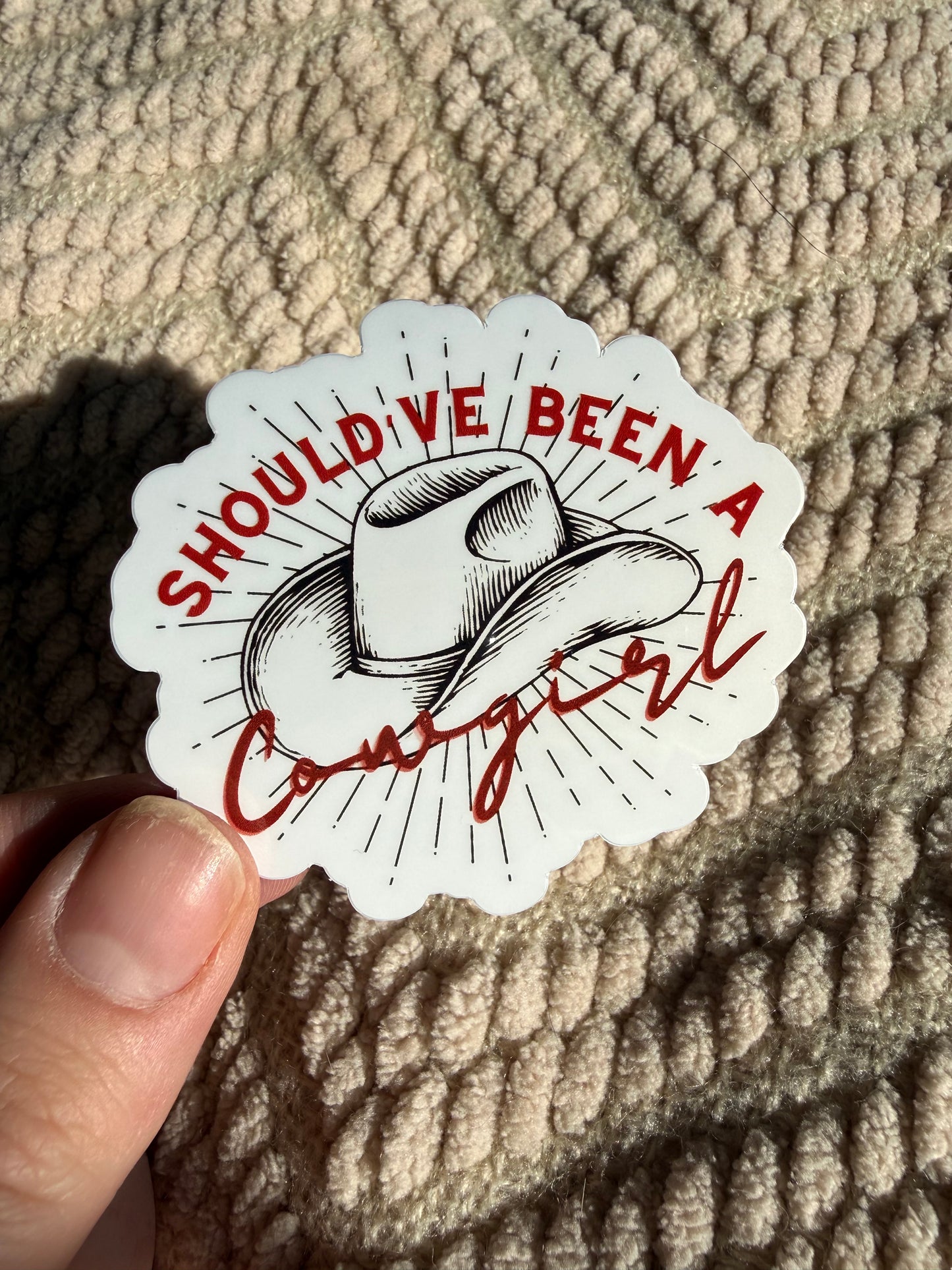 Hand holding "The Wild West Cowgirl Club" sticker featuring a retro cowgirl in a martini glass; chic western aesthetic decal shown in warm sunlight on a tan blanket.