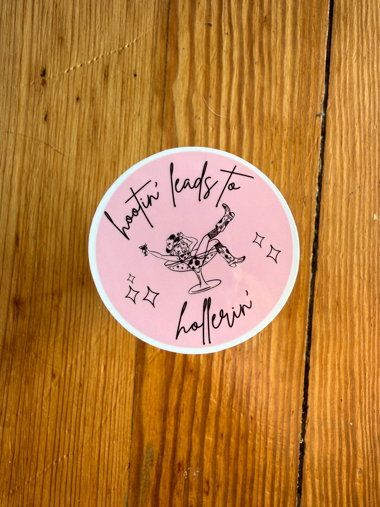 Single 2-inch round pink “Hootin’ Leads to Hollerin’” sticker shown on a wooden background to highlight size and color. This southern humor vinyl sticker is perfect for country girls, western lifestyle fans, and anyone who loves funny country sayings. A great pink western sticker for decorating laptops, notebooks, and as a small country-themed gift or stocking stuffer.