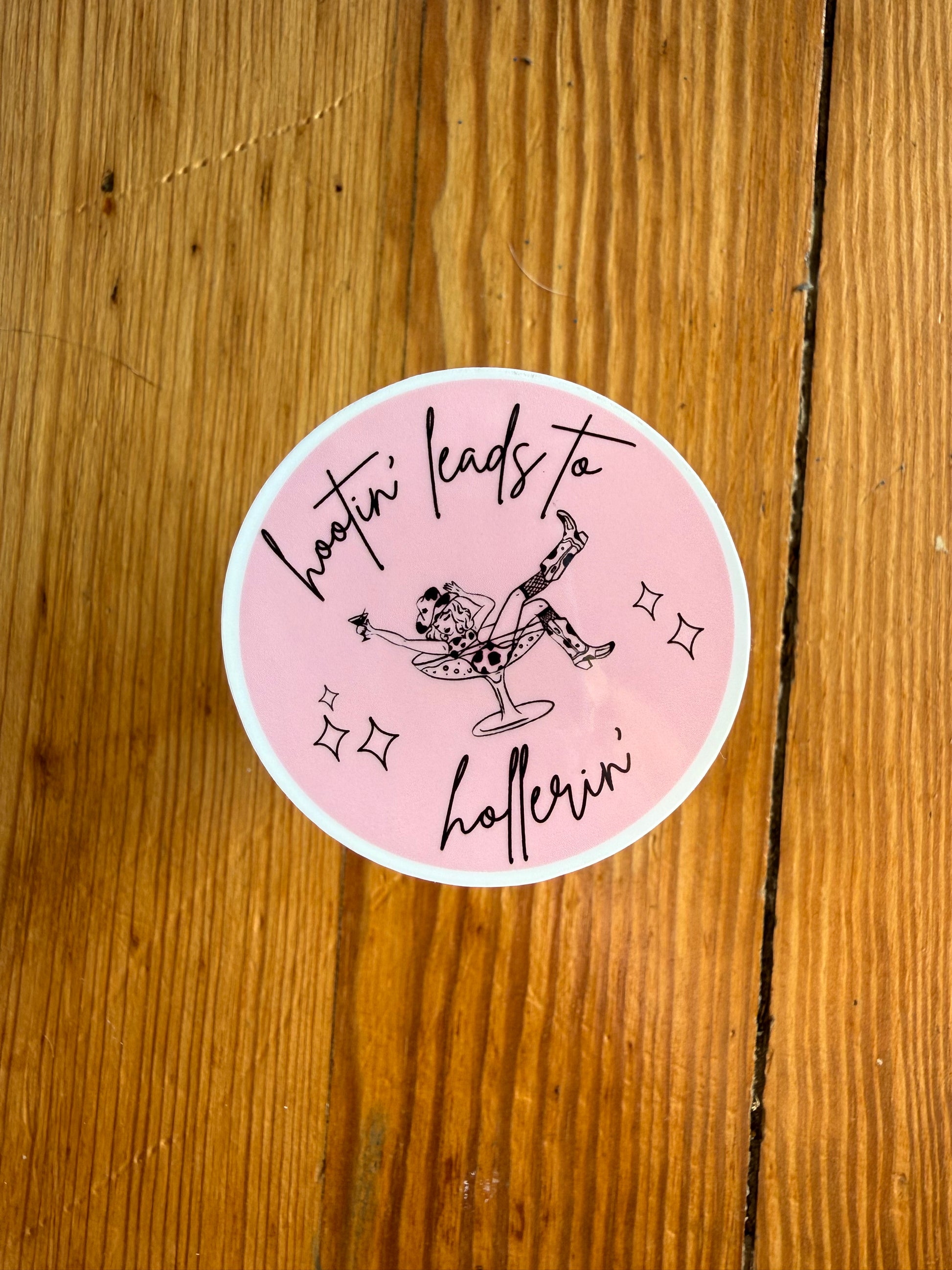 Single 2-inch round pink “Hootin’ Leads to Hollerin’” sticker shown on a wooden background to highlight size and color. This southern humor vinyl sticker is perfect for country girls, western lifestyle fans, and anyone who loves funny country sayings. A great pink western sticker for decorating laptops, notebooks, and as a small country-themed gift or stocking stuffer.