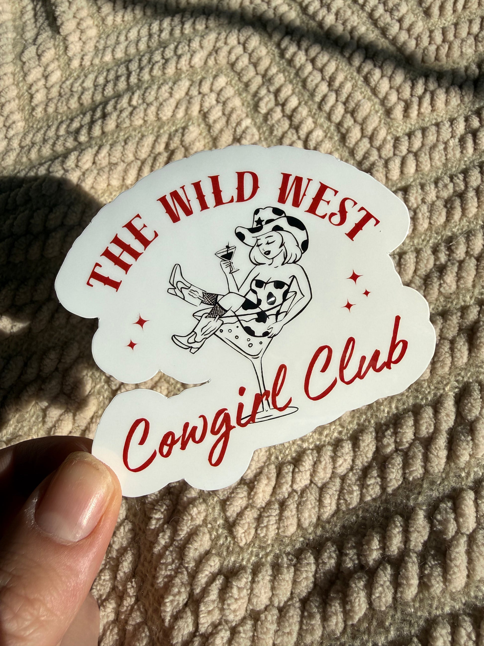 Hand holding "The Wild West Cowgirl Club" sticker featuring a retro cowgirl in a martini glass; chic western aesthetic decal shown in warm sunlight on a tan blanket.