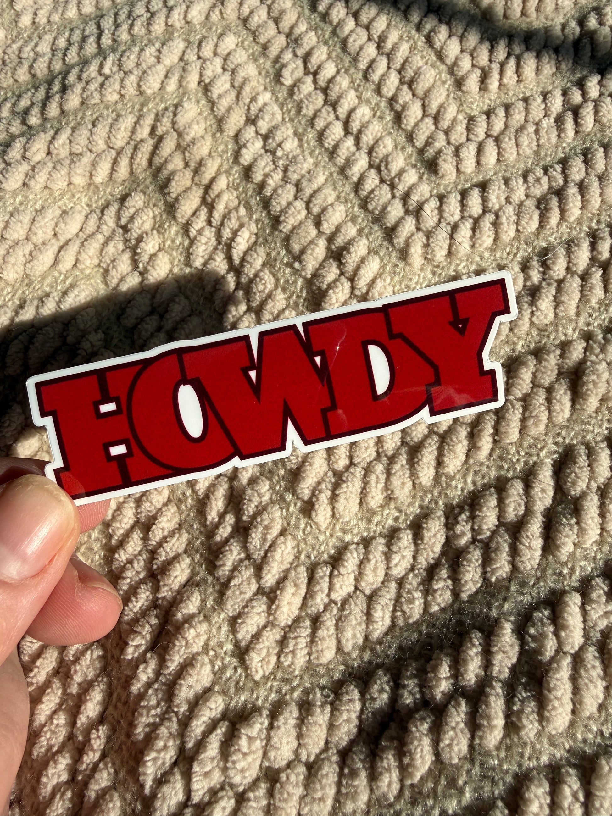 Retro red "Howdy" script sticker being held against a tan blanket; aesthetic western vinyl sticker for boho cowgirl style.
