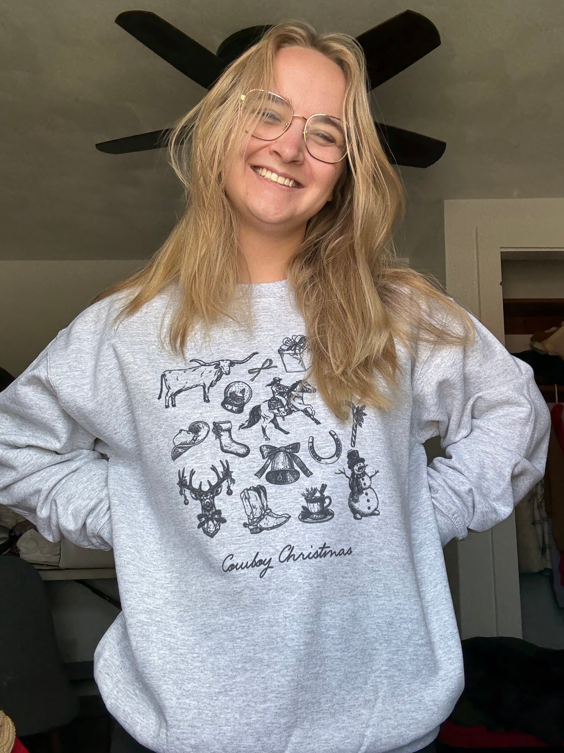 A woman wearing an ash gray cowboy Christmas crewneck with a vintage Western holiday collage featuring retro cowboy sketches, Christmas trees, stars, lassos, and rustic country illustrations. Cozy Western Christmas sweatshirt styled with jeans, perfect for cowgirl Christmas outfits, holiday rodeo gifts, and rustic farmhouse winter apparel.