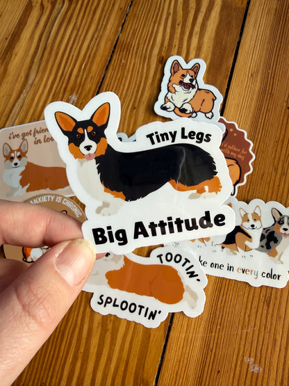 "Tiny Legs, Big Attitude": A hand holds a sticker featuring a sassy corgi graphic with the phrase "Tiny legs, big attitude."