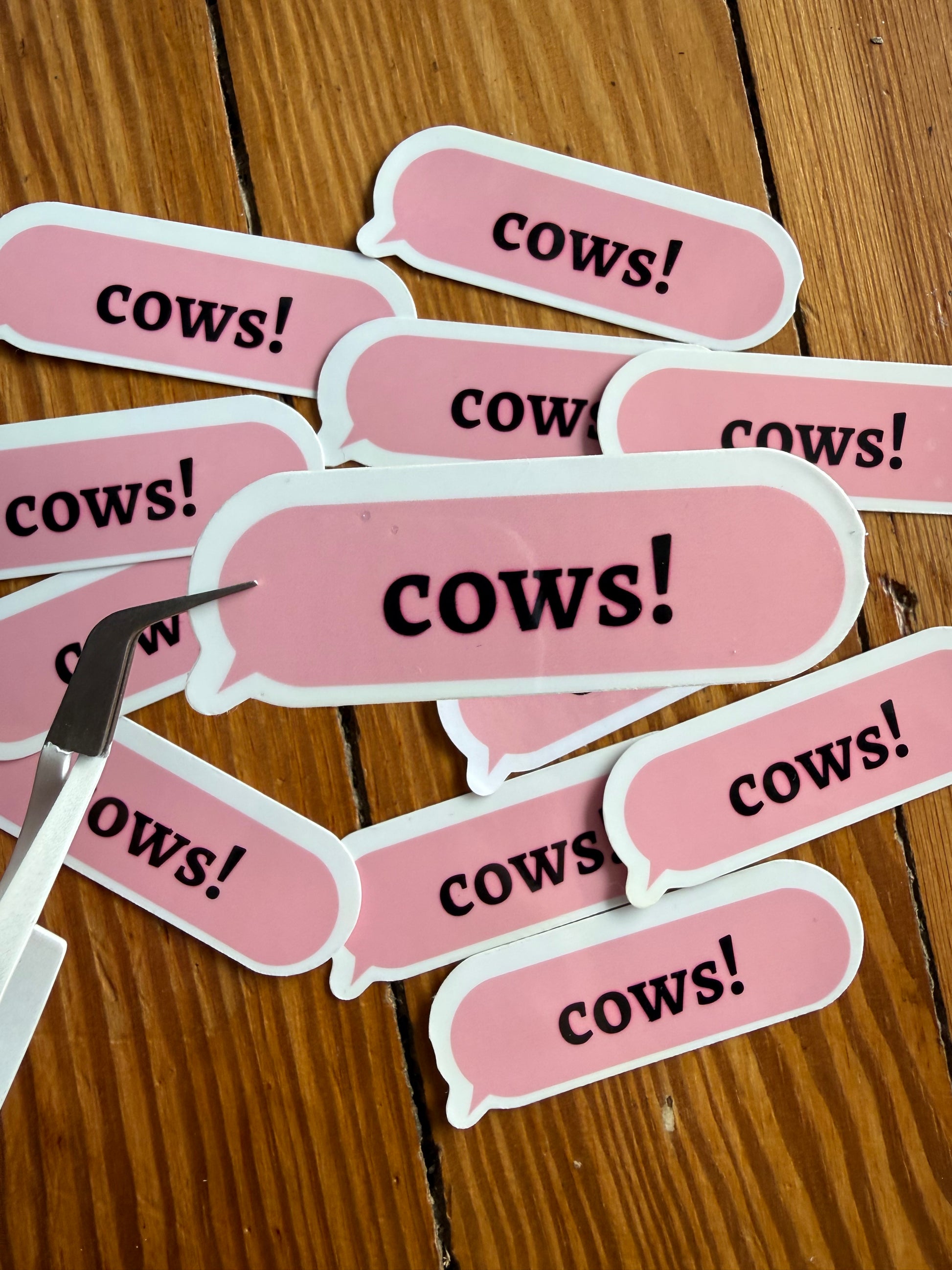 Close-up of silver craft tweezers holding one pink "Cows!" speech bubble sticker over a pile of stickers, emphasizing the high-quality vinyl material and precise cut for planners and journaling.