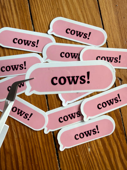Close-up of silver craft tweezers holding one pink "Cows!" speech bubble sticker over a pile of stickers, emphasizing the high-quality vinyl material and precise cut for planners and journaling.