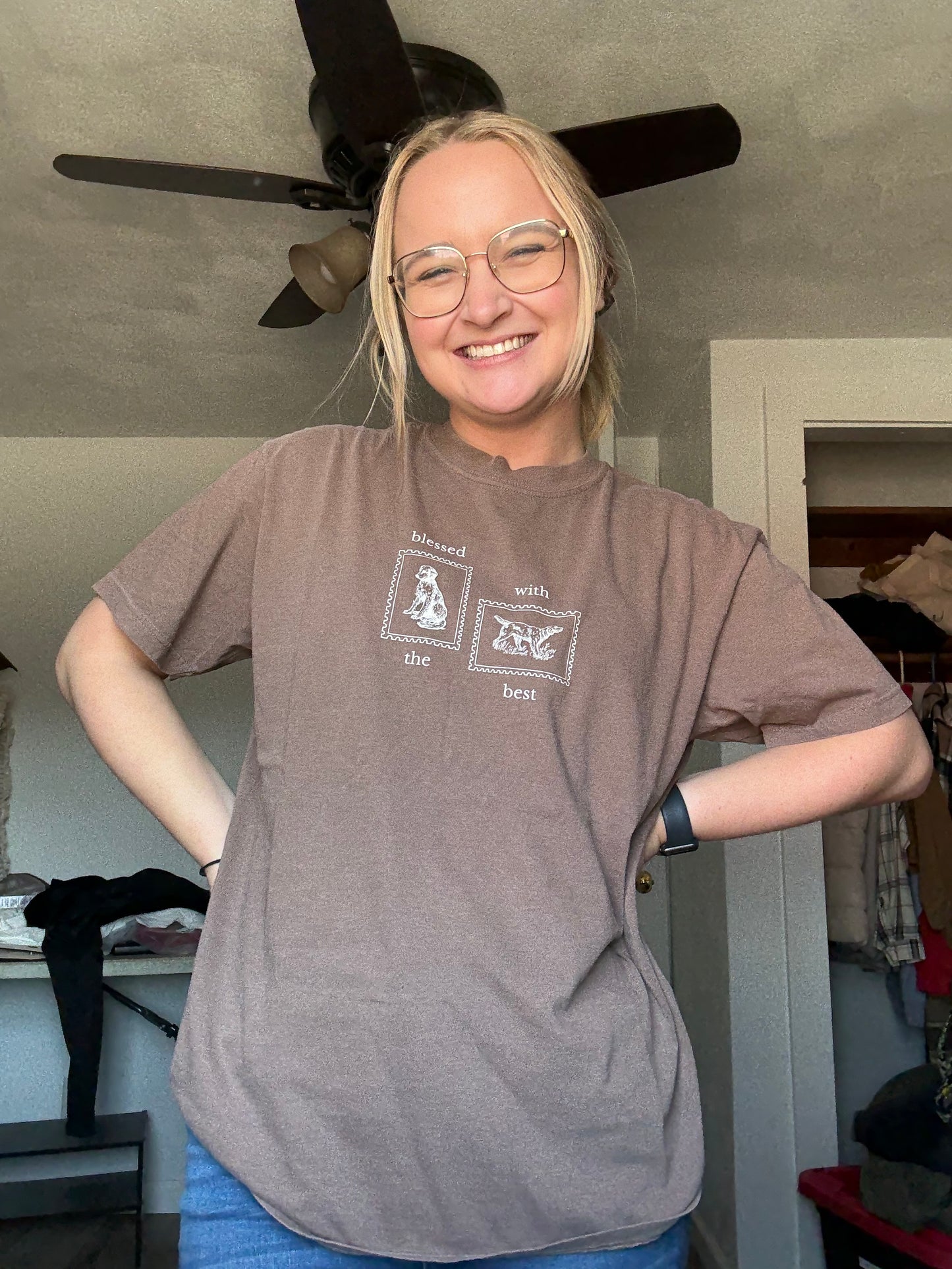 Person wearing a brown t-shirt tan Blessed with the Best text and graphic of two stamps with dogs on them, standing indoors.