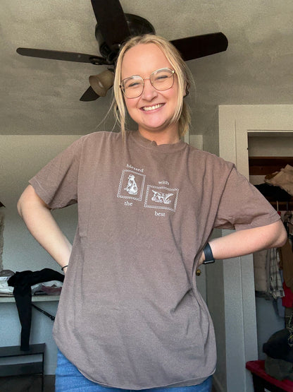 Person wearing a brown t-shirt tan Blessed with the Best text and graphic of two stamps with dogs on them, standing indoors.