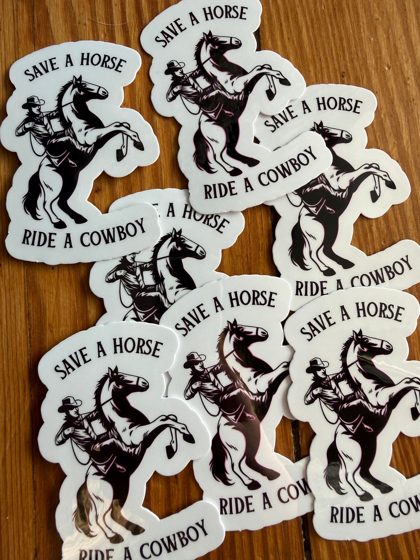 A grouping of “Save a Horse, Ride a Cowboy” vinyl stickers arranged on a wooden background, showcasing the full design and finish. This western sticker features a classic cowboy riding a rearing horse with a humorous country phrase. Great for western decor lovers, cowgirl and cowboy gifts, southern humor stickers, rodeo fans, 90s country music lovers and country-themed vinyl decals for laptops, notebooks, and planners.