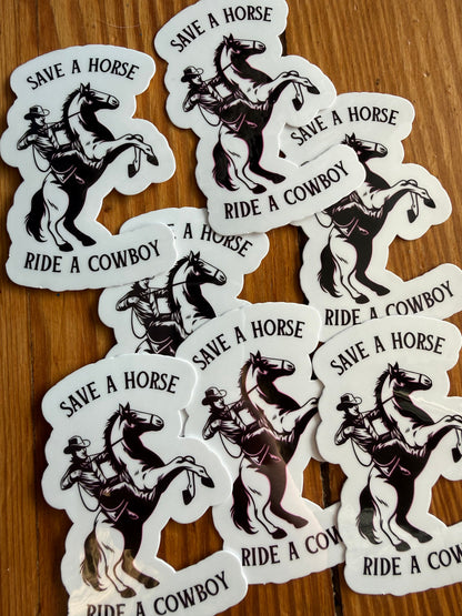 A grouping of “Save a Horse, Ride a Cowboy” vinyl stickers arranged on a wooden background, showcasing the full design and finish. This western sticker features a classic cowboy riding a rearing horse with a humorous country phrase. Great for western decor lovers, cowgirl and cowboy gifts, southern humor stickers, rodeo fans, 90s country music lovers and country-themed vinyl decals for laptops, notebooks, and planners.